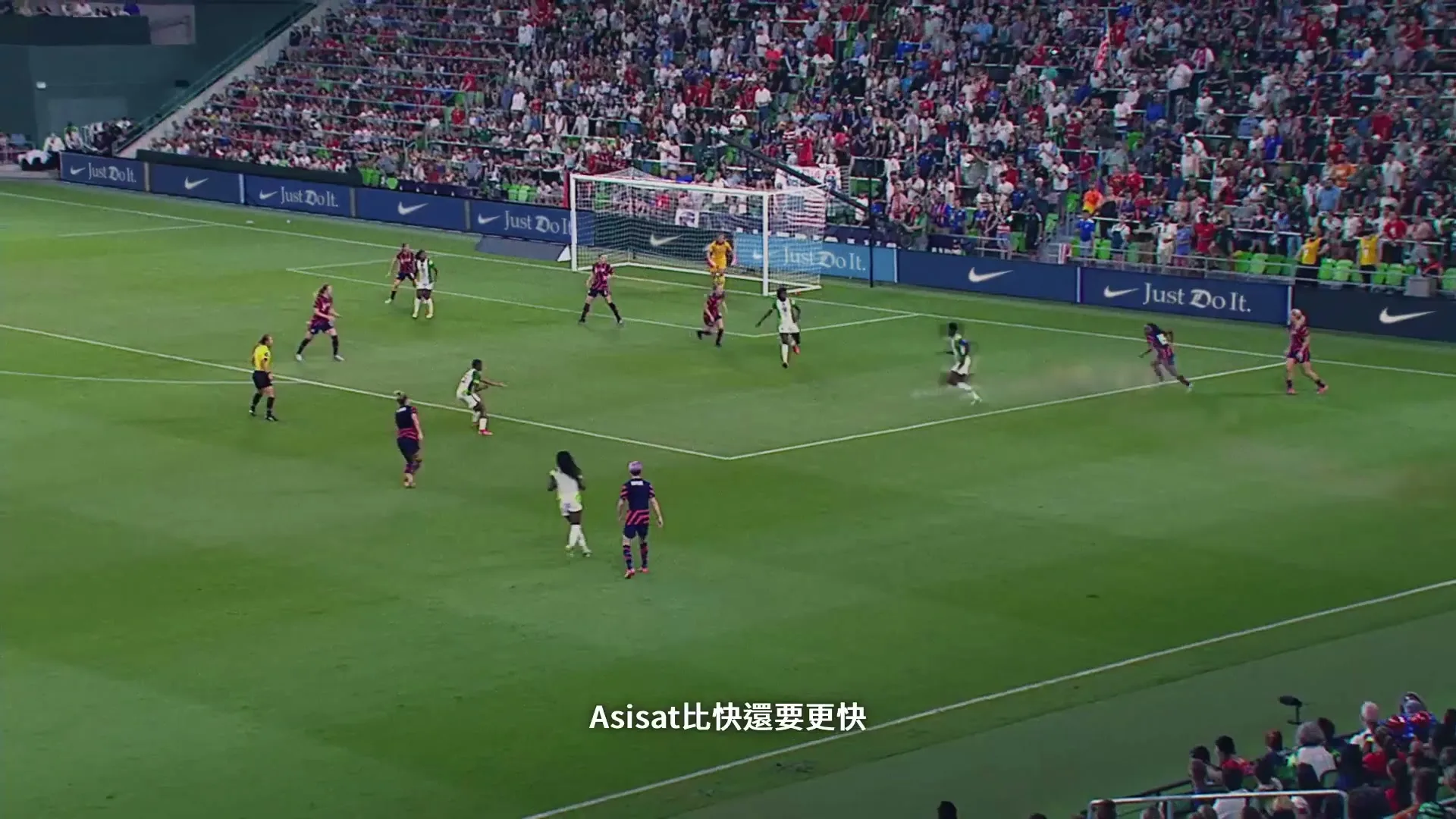A wide shot of a women's soccer match in a stadium, with players in white and red uniforms on the field and spectators in the stands.