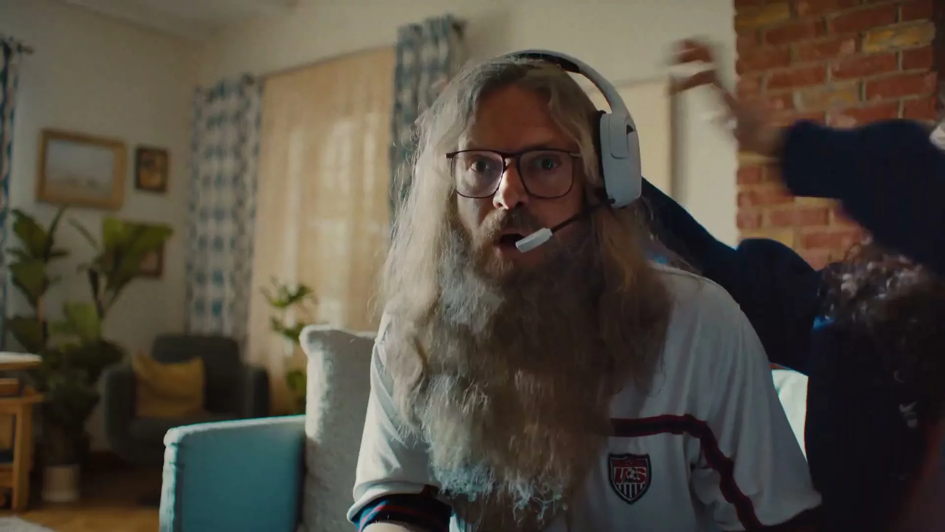 A man with long hair and beard wears a headset and glasses, looking surprised while sitting on a couch in a living room.
