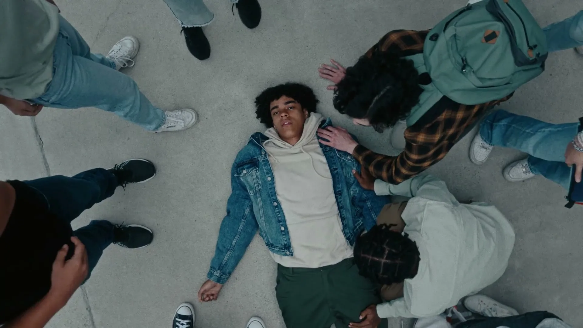 A young man lies on the ground, surrounded by a group of concerned teenagers, as one tries to help him up in an outdoor setting.