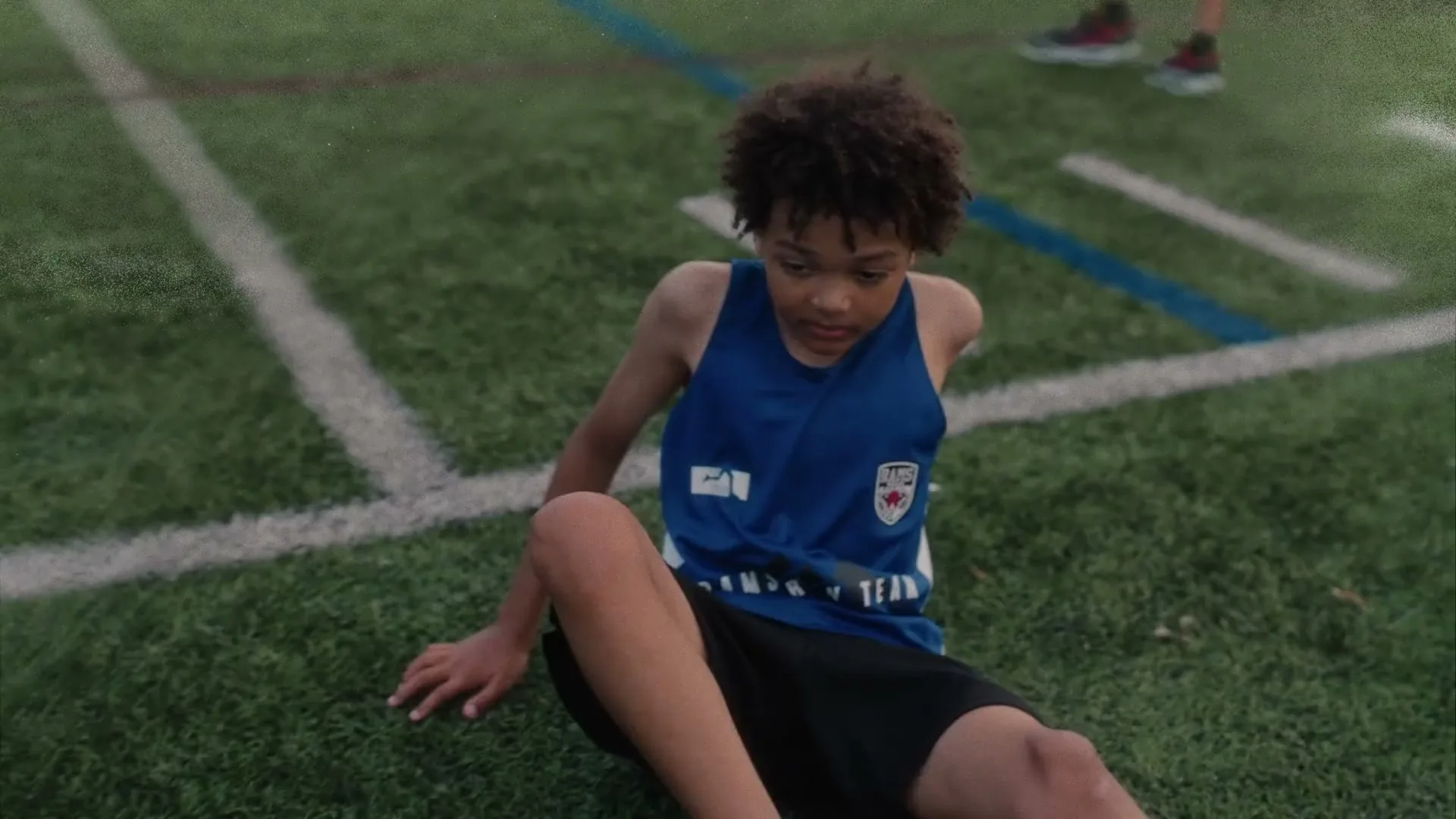 A young boy with curly hair, wearing a blue and white athletic tank top and black shorts, sits on a green sports field with white and blue lines, looking down with a somber expression.
