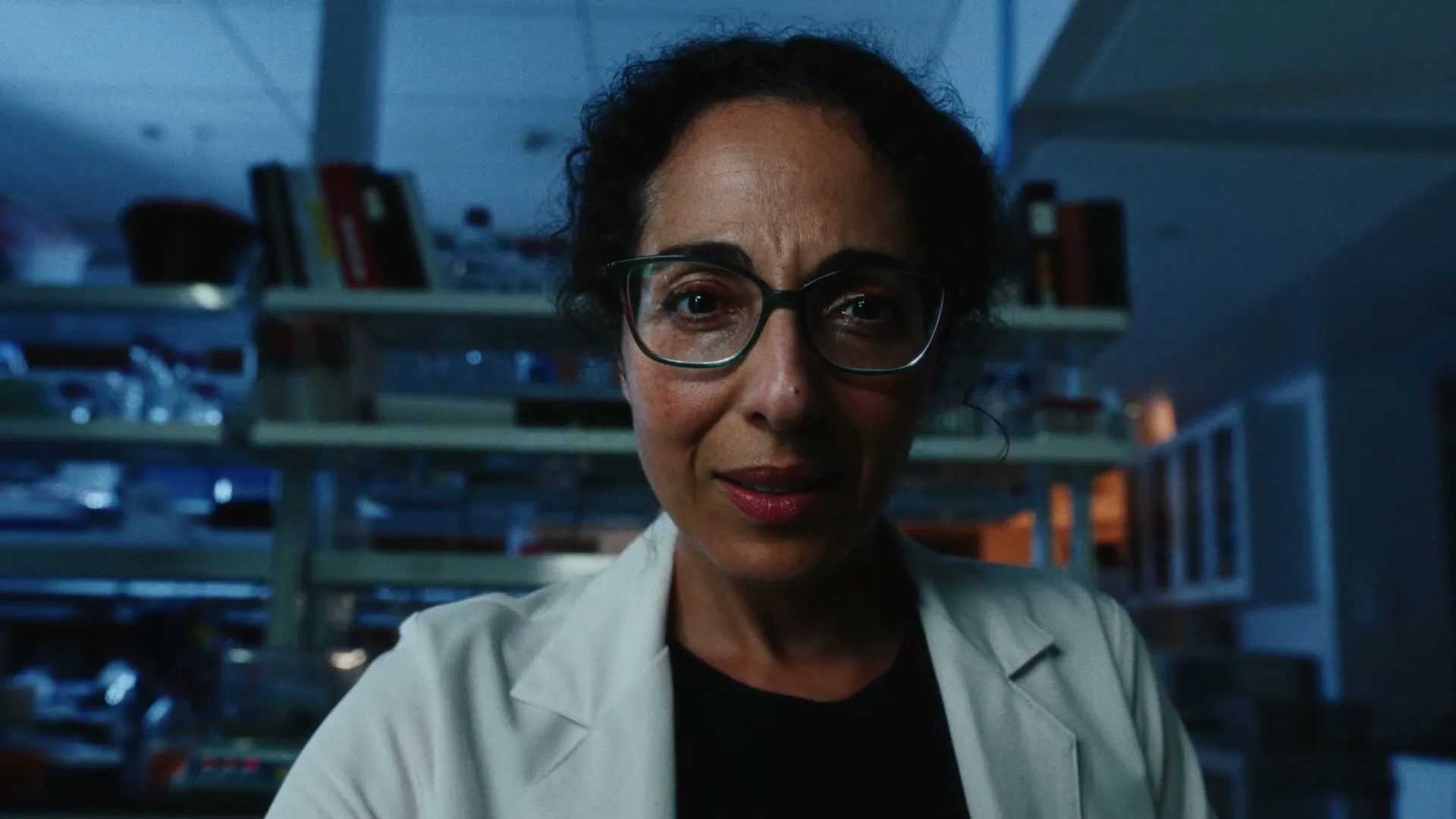 A female scientist in a lab coat and glasses looks directly at the camera in a dimly lit laboratory.