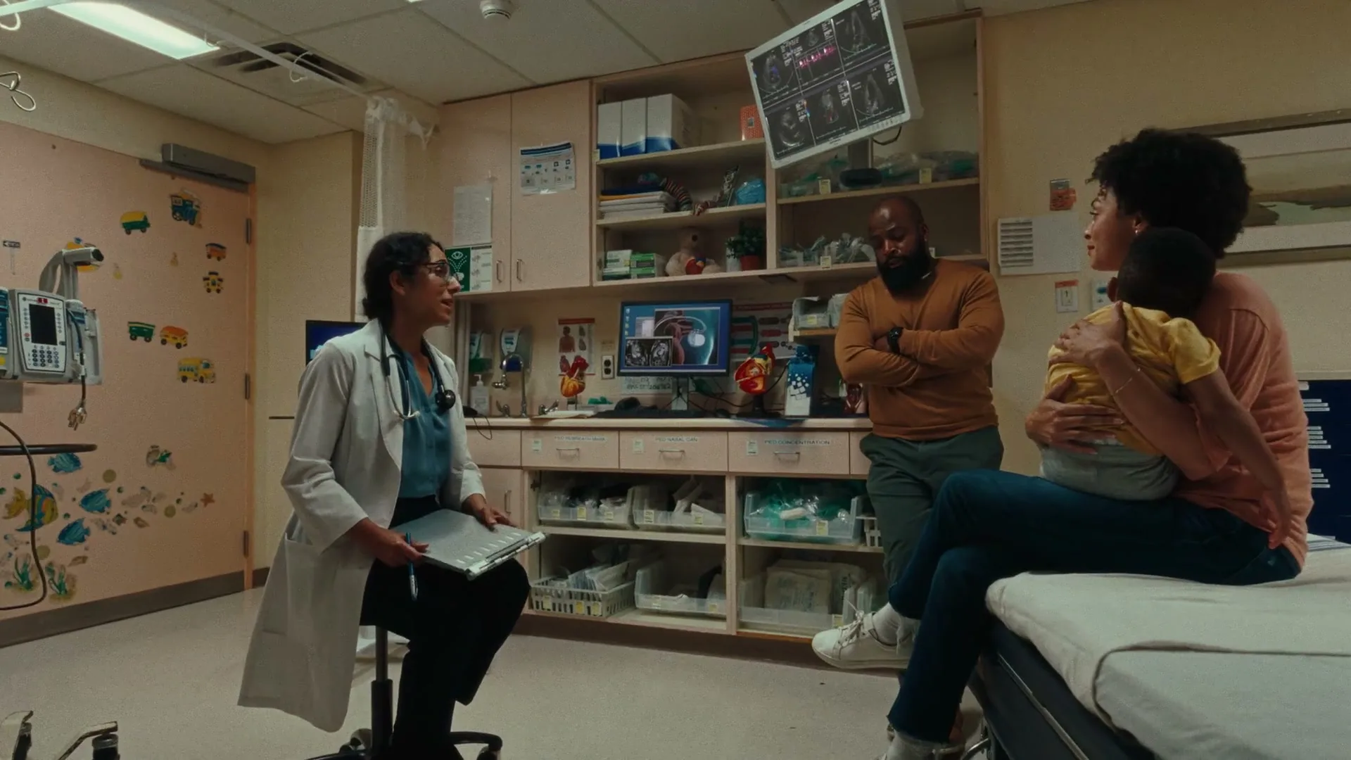 A female doctor in a white coat talks to a couple with a baby in a hospital room, discussing medical information on a screen.