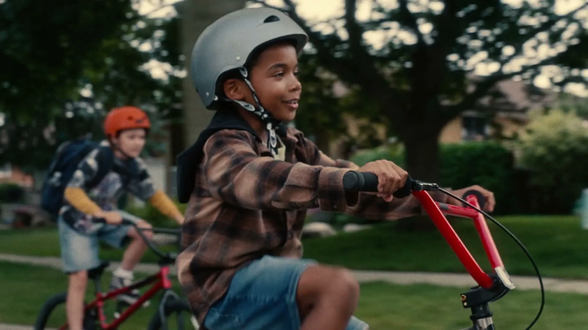 Two young boys, one in a grey helmet and plaid shirt, the other in an orange helmet and patterned shirt, ride bicycles down a tree-lined suburban street.