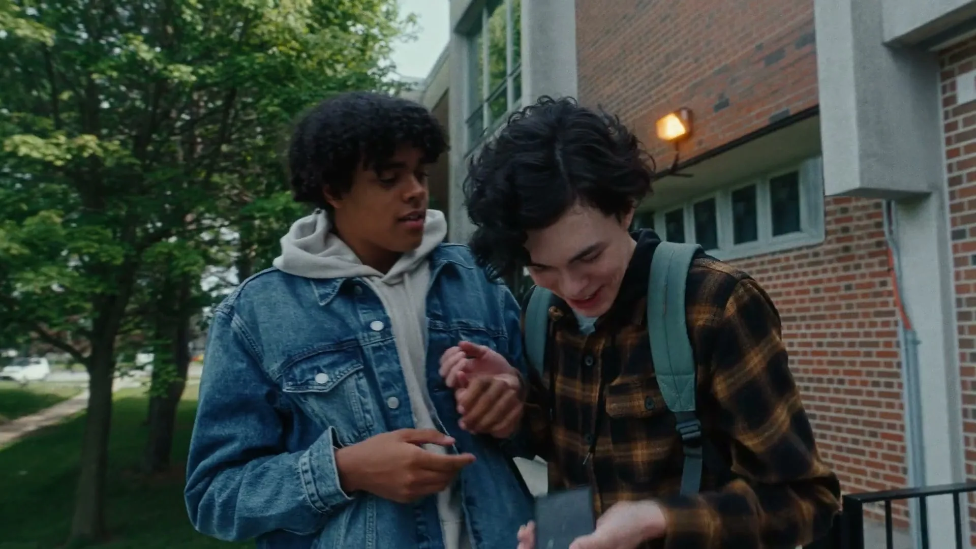 Two male teens, one with curly hair and a denim jacket, the other with a plaid shirt and backpack, look at a phone and laugh outside a brick building.