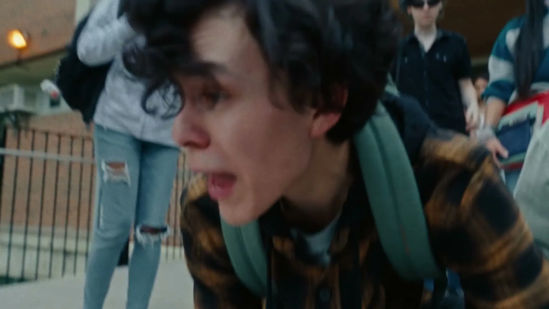 A male teen with curly hair and a backpack looks shocked and concerned while kneeling over someone on the ground outside a school.