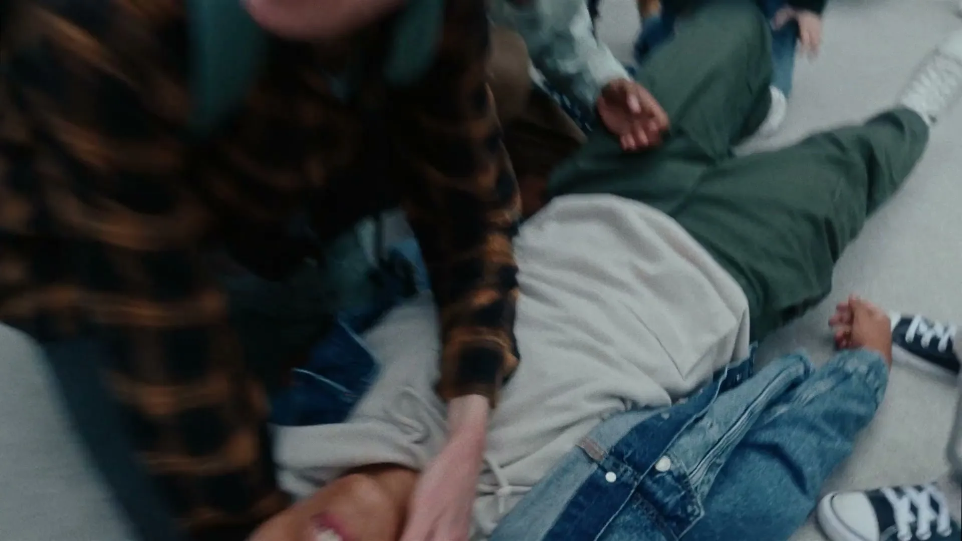 A male teen in a plaid shirt kneels over another male teen lying on the floor, grabbing his face, in a school hallway.