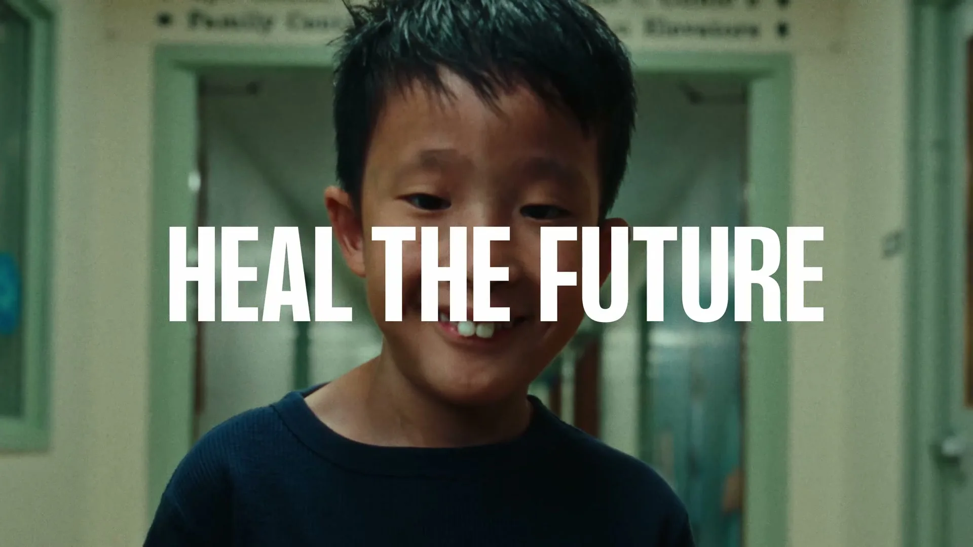 A young Asian boy with a gap-toothed smile looks directly at the camera in a hospital hallway, with the text "HEAL THE FUTURE" superimposed.