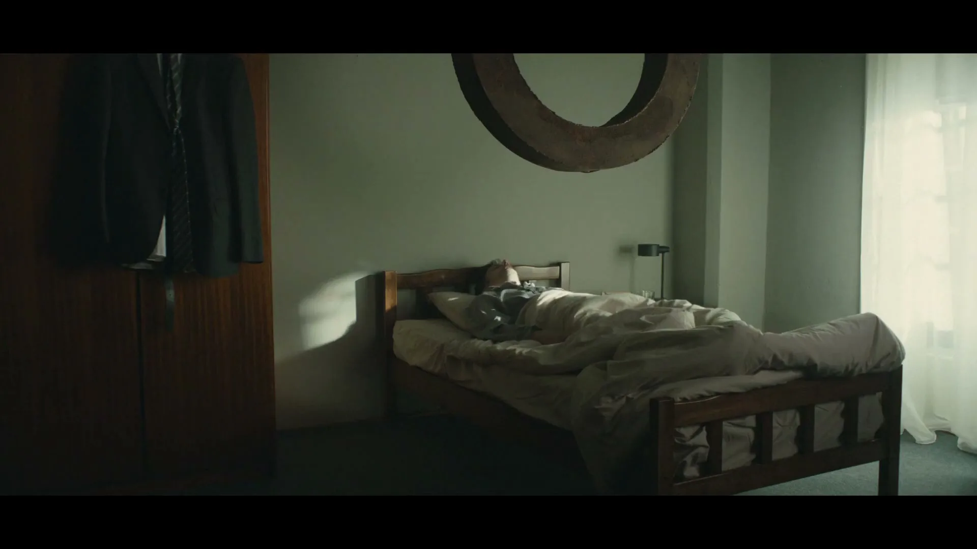 A man wakes up in bed with a large, heavy ring hovering over his head in a dimly lit bedroom.
