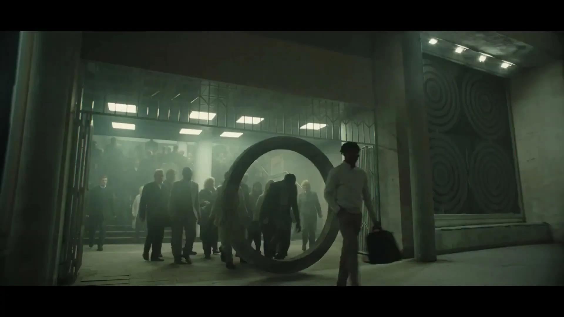 A diverse group of people in business attire walk through a large, dark, industrial-looking building, with one man pushing a large metal ring.