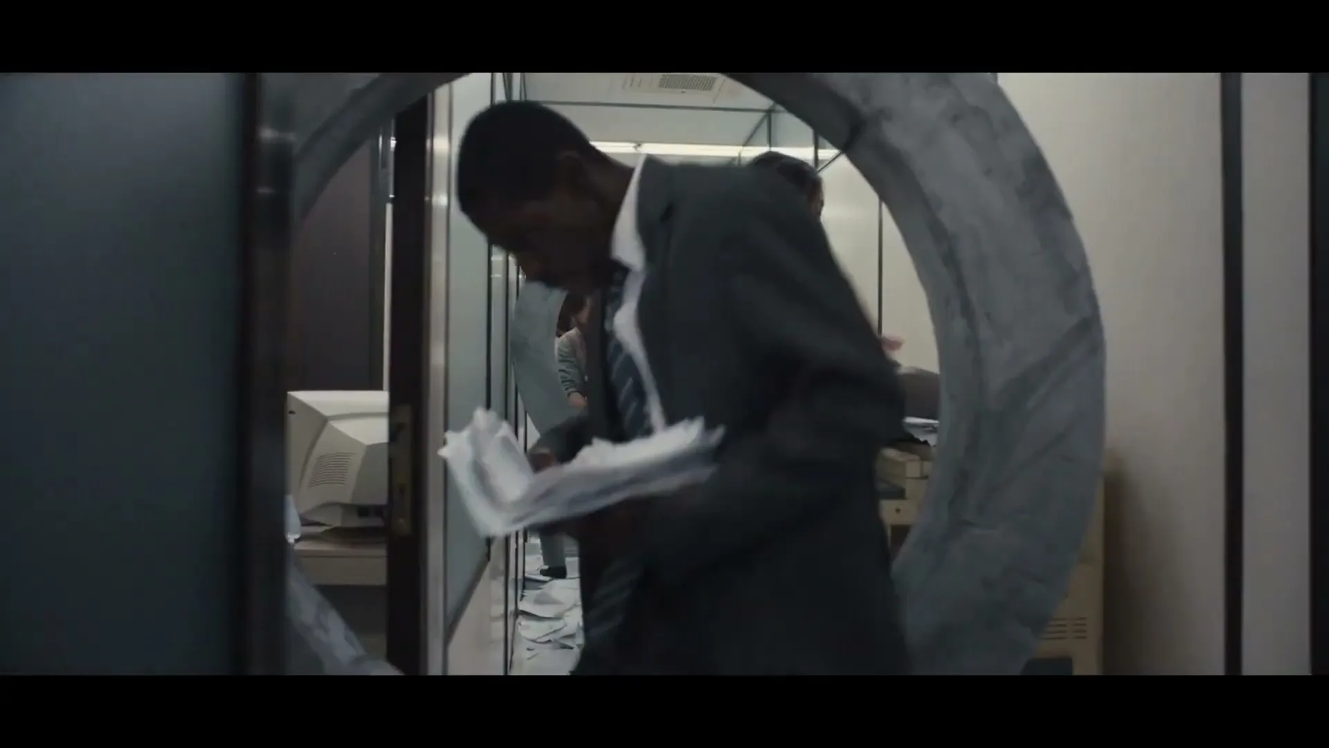 A diverse group of office workers in business attire navigate a chaotic office, jumping through concrete hoops and carrying stacks of paper.