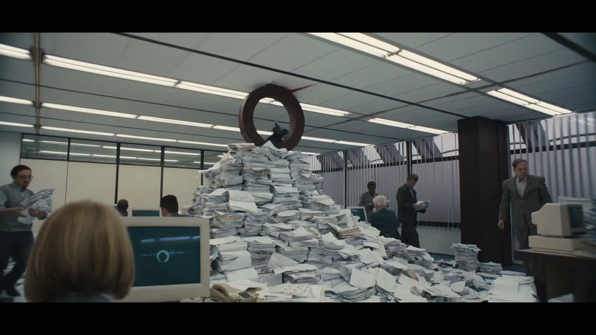 An office worker sits at a desk, looking at a computer screen, while a large pile of papers with a hoop on top dominates the center of a busy office.