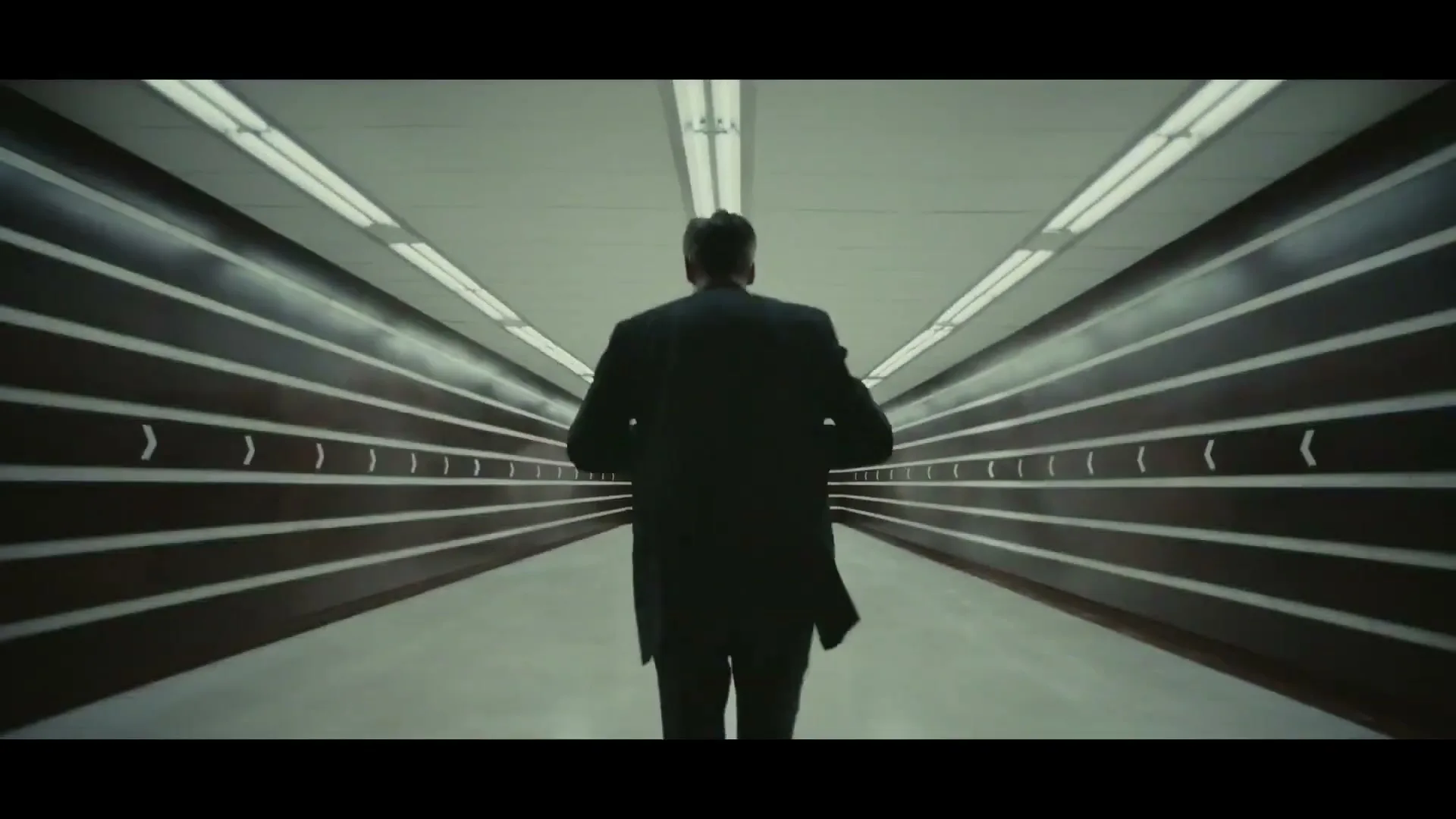A man in a suit runs away from the camera down a long, brightly lit, futuristic tunnel with horizontal stripes on the walls.