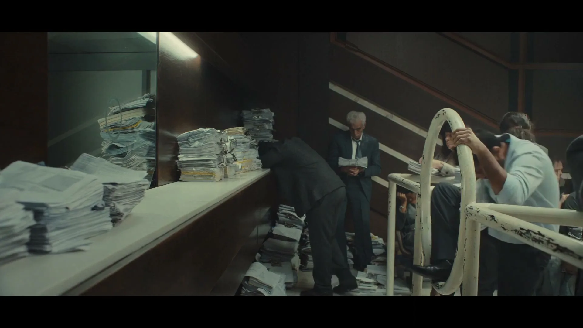 A group of people are seen in a dimly lit, cluttered office space, surrounded by stacks of paper, as they navigate a staircase.