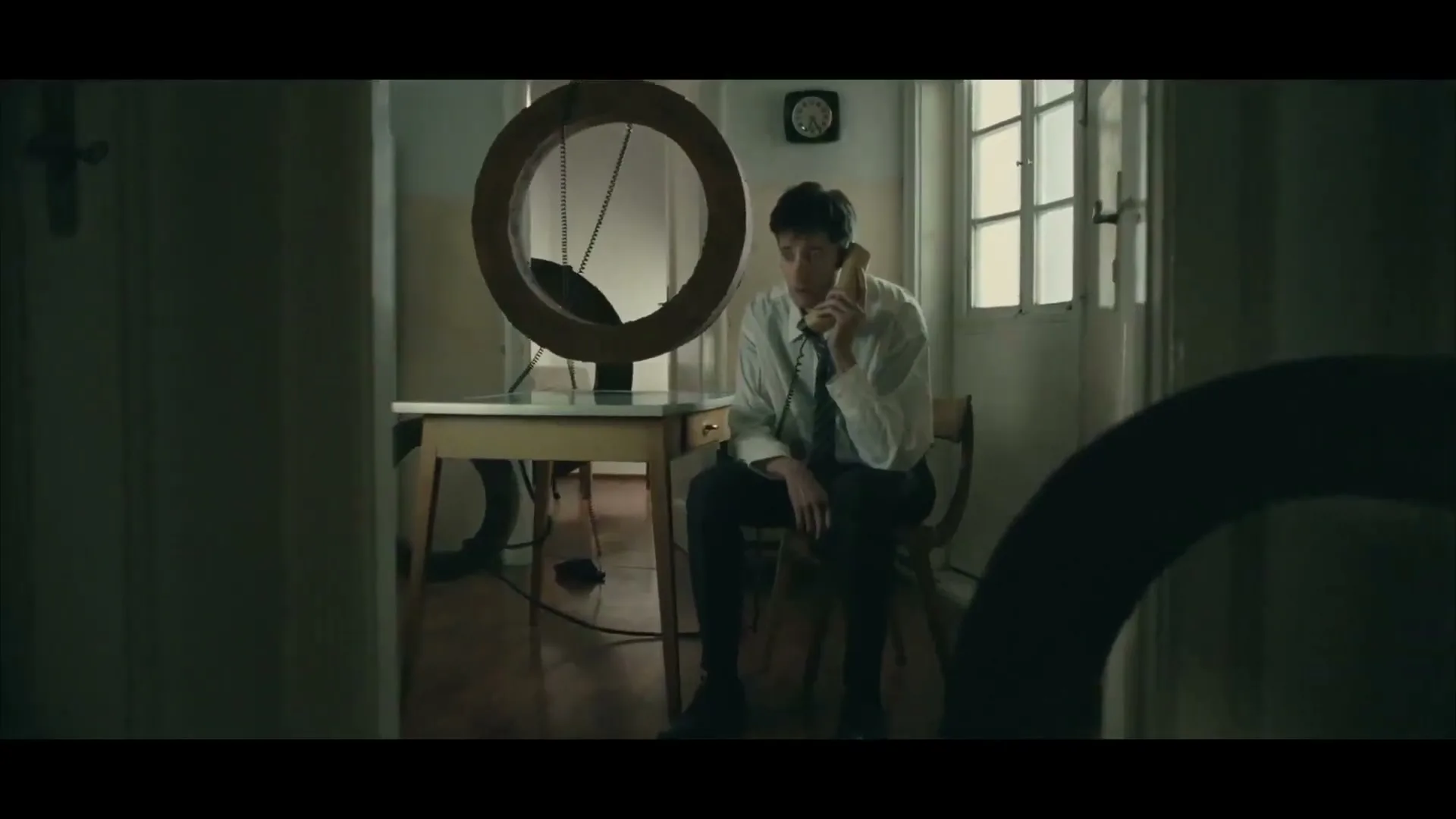 A young man in a white shirt and tie sits at a desk, holding a phone to his ear, looking confused in a dimly lit room.