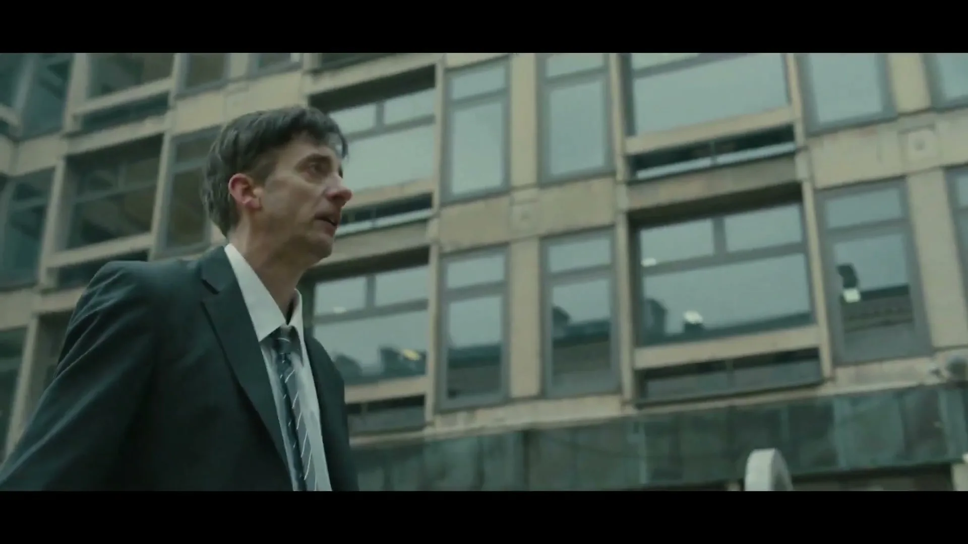A man in a suit walks past a building, then looks up at a metal structure, appearing confused or surprised.
