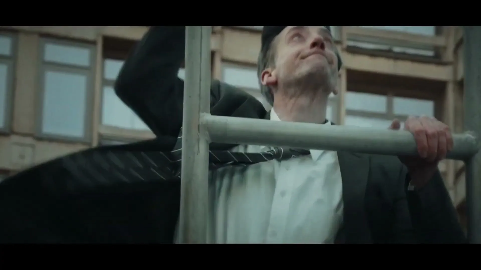 A man in a suit struggles to pull himself up a metal ladder, with a building in the background.