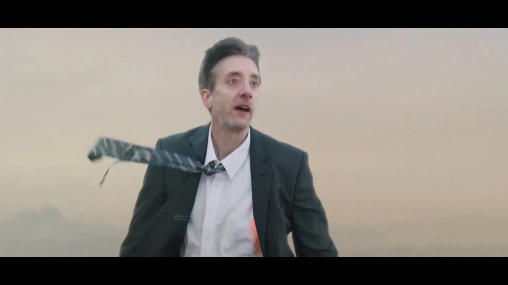 A man in a suit and tie, with his hair and tie blowing in the wind, looks around with a surprised expression against a cloudy sky.