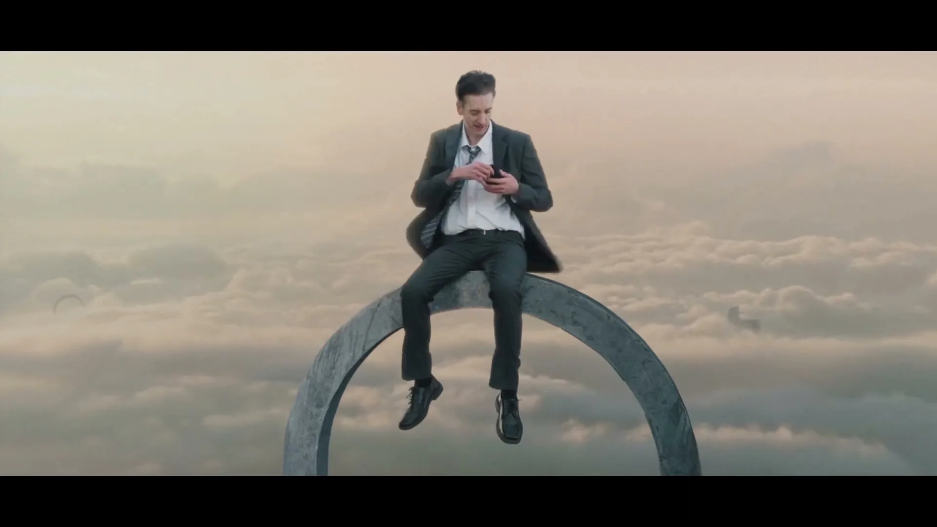 A young man in a suit sits on a large metal ring above the clouds, pulling out his phone and looking at it.