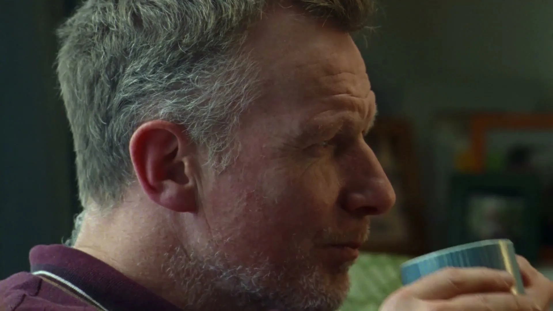 A middle-aged man with grey hair and stubble sips from a blue mug, looking off-screen in a domestic interior.
