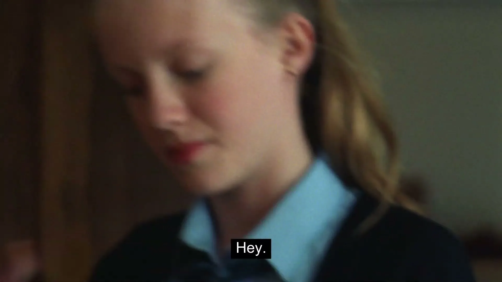 A young blonde girl in a school uniform looks down, then turns her head to the right, as if someone is calling her name.