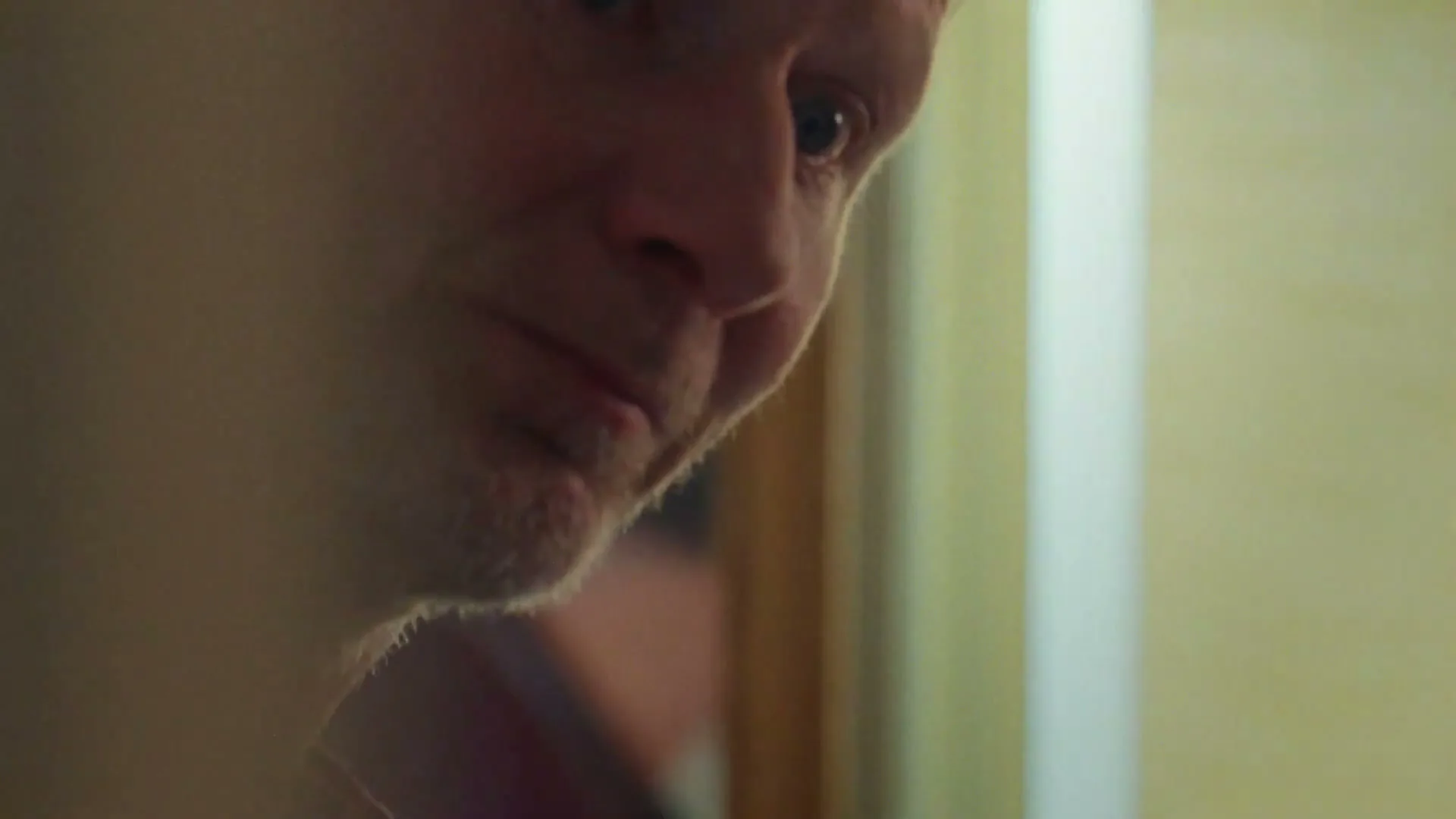 A man with short hair and stubble looks down, then up with a slight smile, in a softly lit interior.