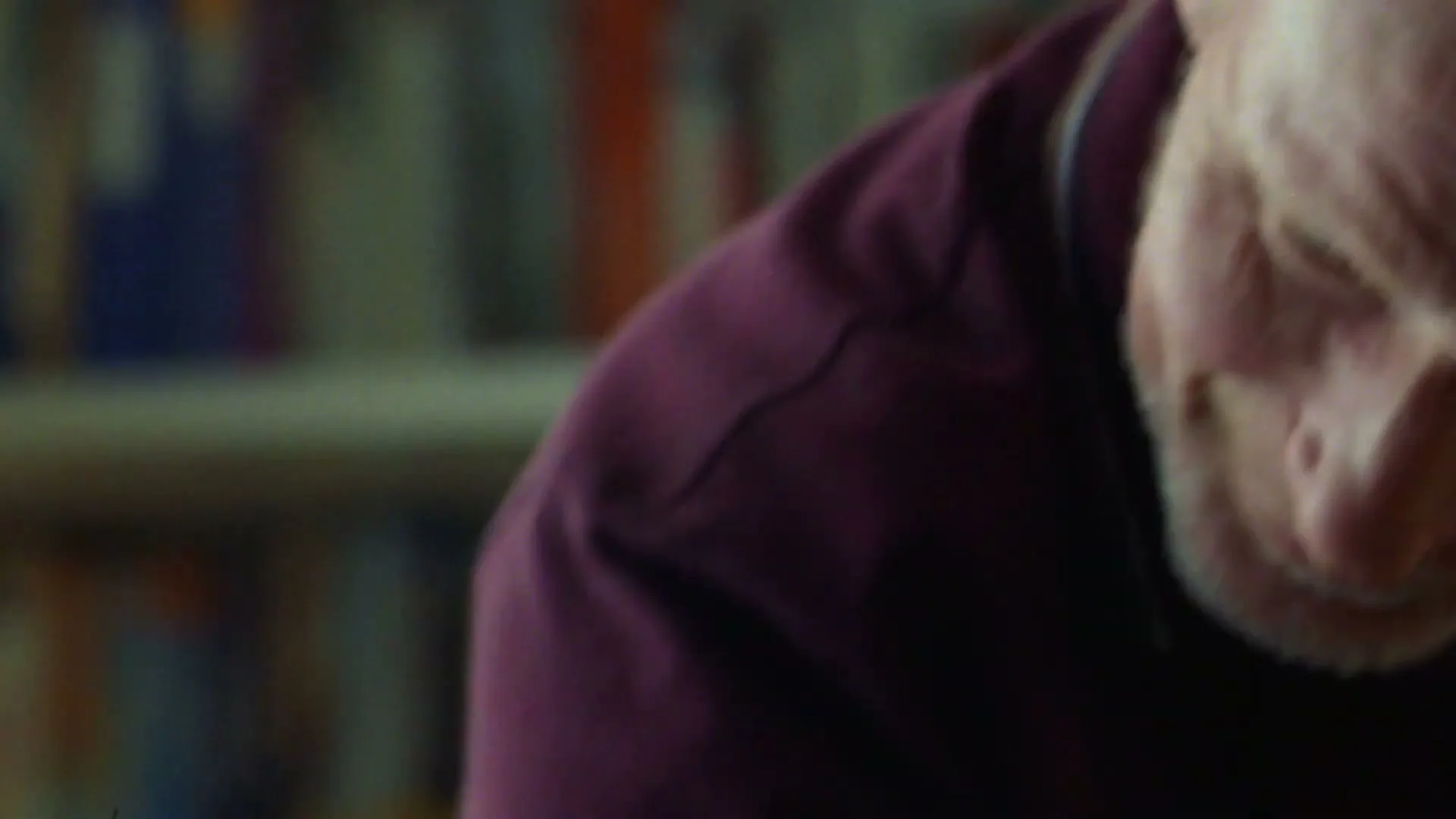 A man with a white beard and purple shirt looks down, then smiles, in an interior setting with blurred bookshelves in the background.