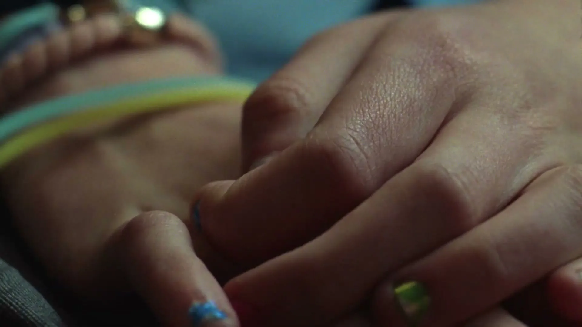 A closeup shot shows two hands, one resting gently on top of the other, with colorful nail polish and bracelets visible.