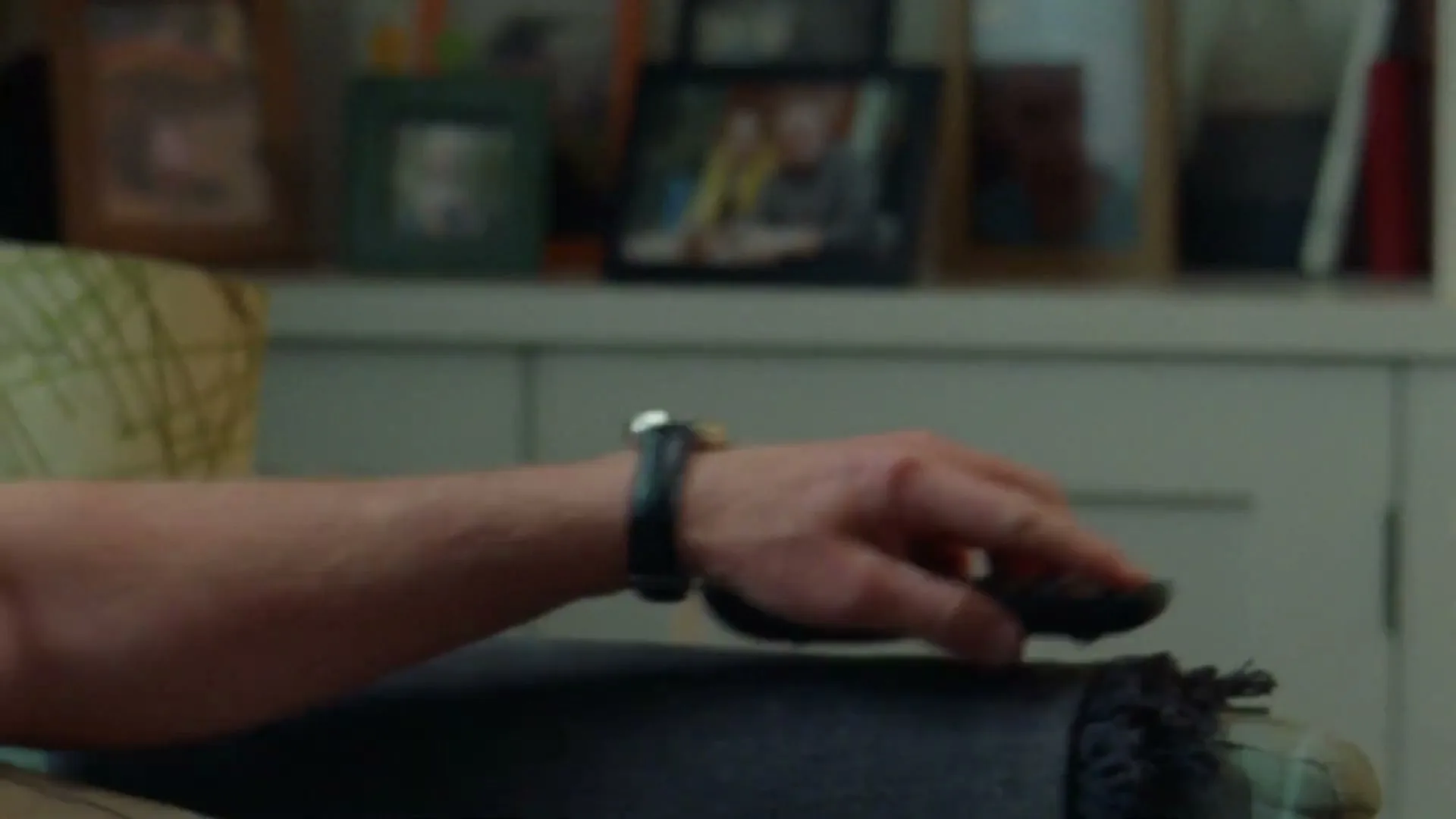 A man's arm and hand, wearing a watch, reaches for a remote control on a blanket, with blurred framed photos in the background.