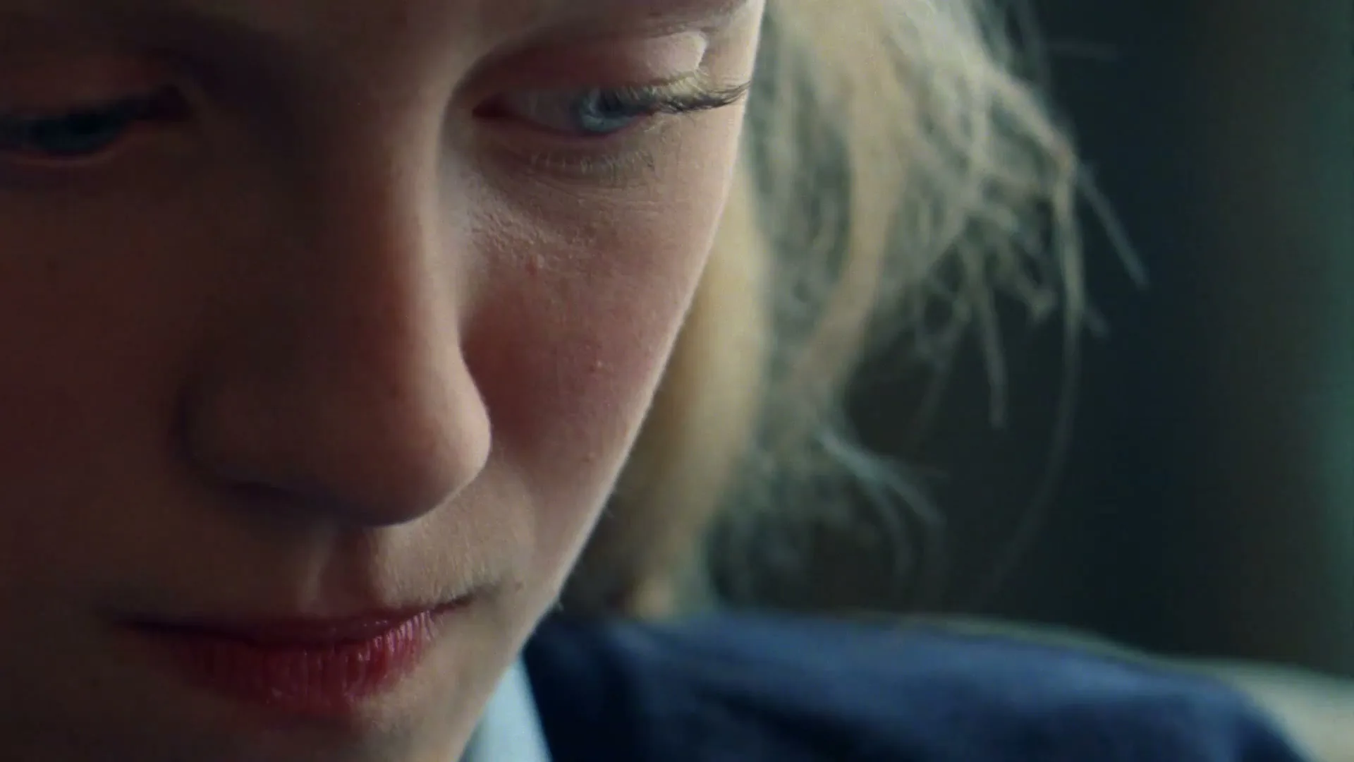 A young blonde woman with blue eyes and red lips looks down with a thoughtful expression, her face partially shadowed.