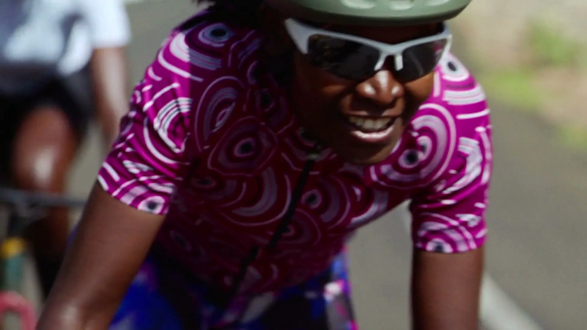 A Black woman in her 30s, wearing a pink patterned cycling jersey, white sunglasses, and a green helmet, smiles while cycling outdoors on a sunny day.