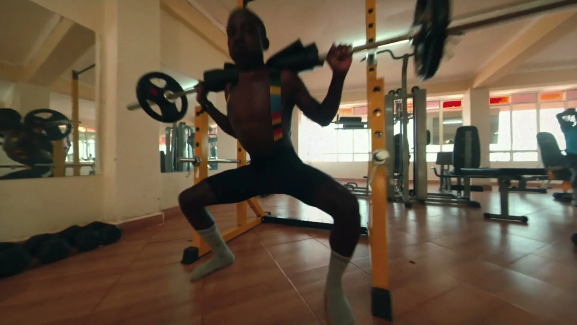 A young man in a black singlet and grey socks performs squats with a barbell in a brightly lit gym.