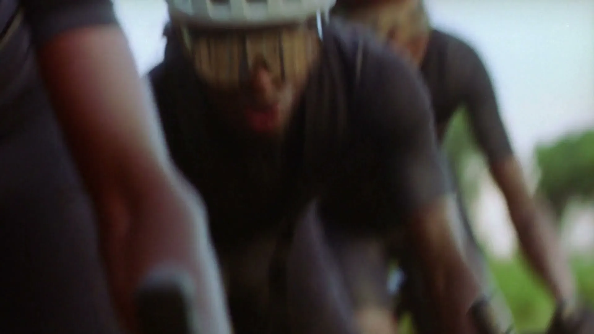 A blurred shot of several cyclists in black gear and helmets, racing outdoors on a bright day.