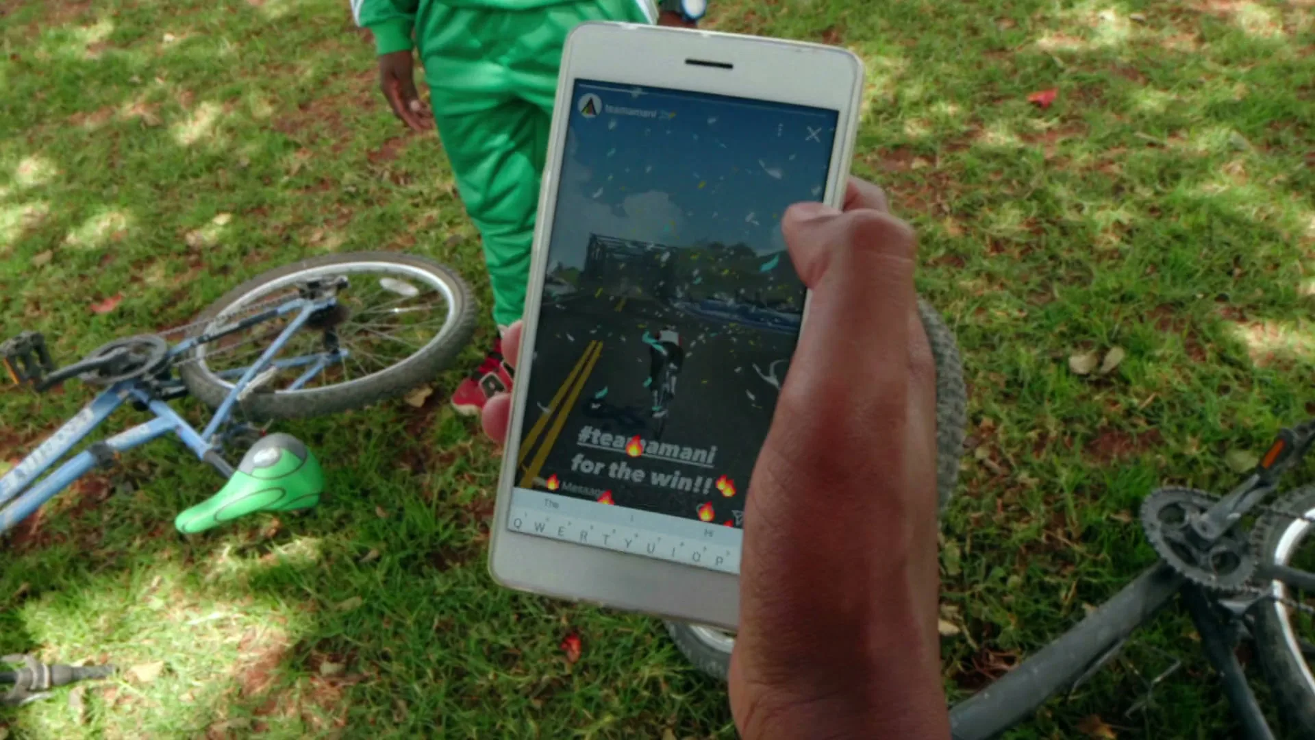 A person's hand holds a white smartphone, displaying a social media interface with a cycling video and text, while a child in a green tracksuit stands nearby on a grassy field with bicycles.