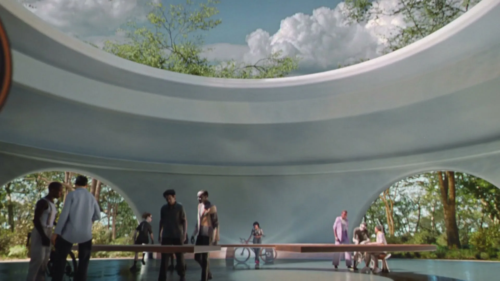 A diverse group of people of all ages interact in a futuristic, open-air circular building with trees and sky visible through large archways.