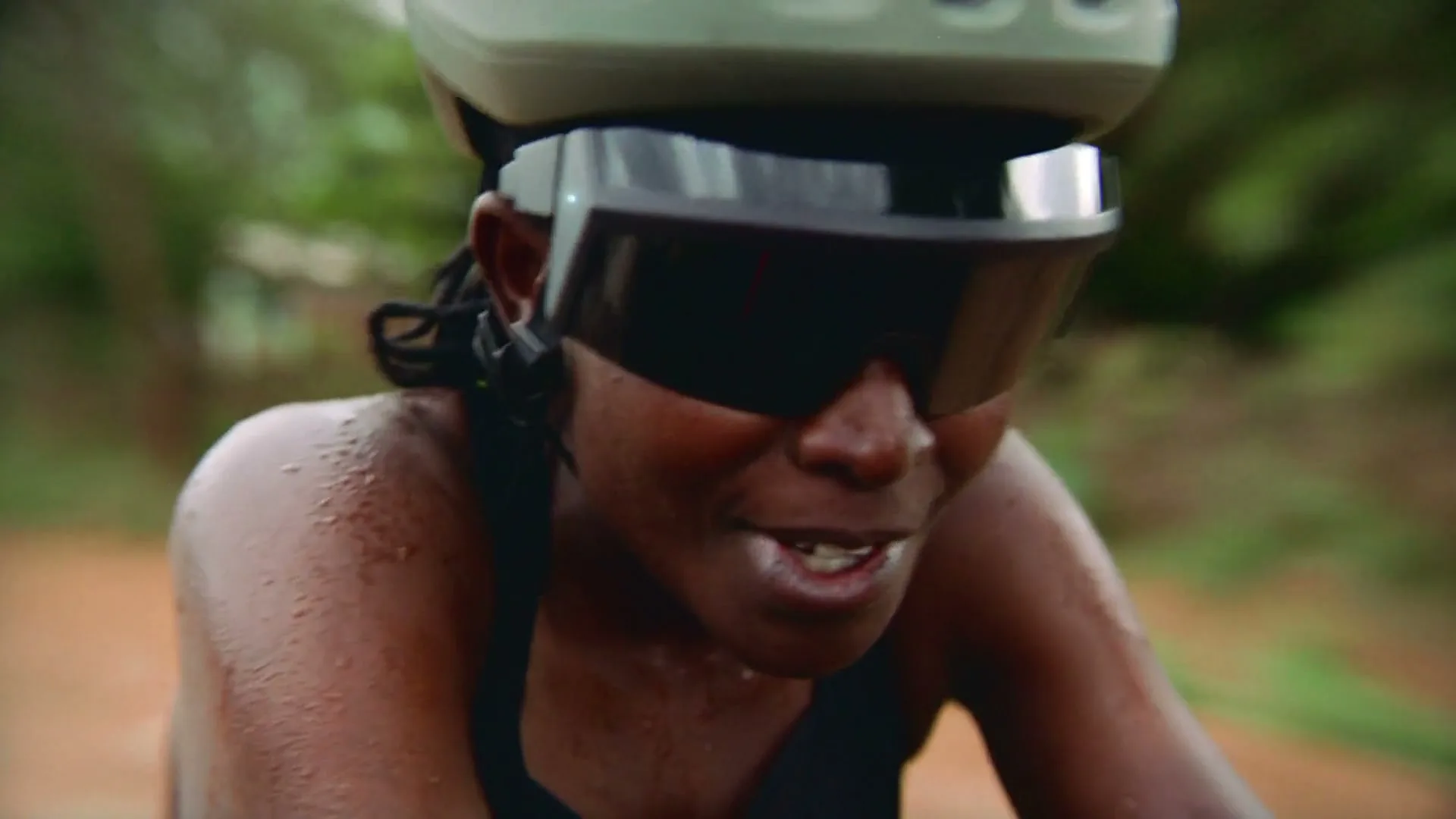 A Black female cyclist, wearing a helmet and sunglasses, is seen sweating and exerting herself while riding a bike outdoors on a dirt path.