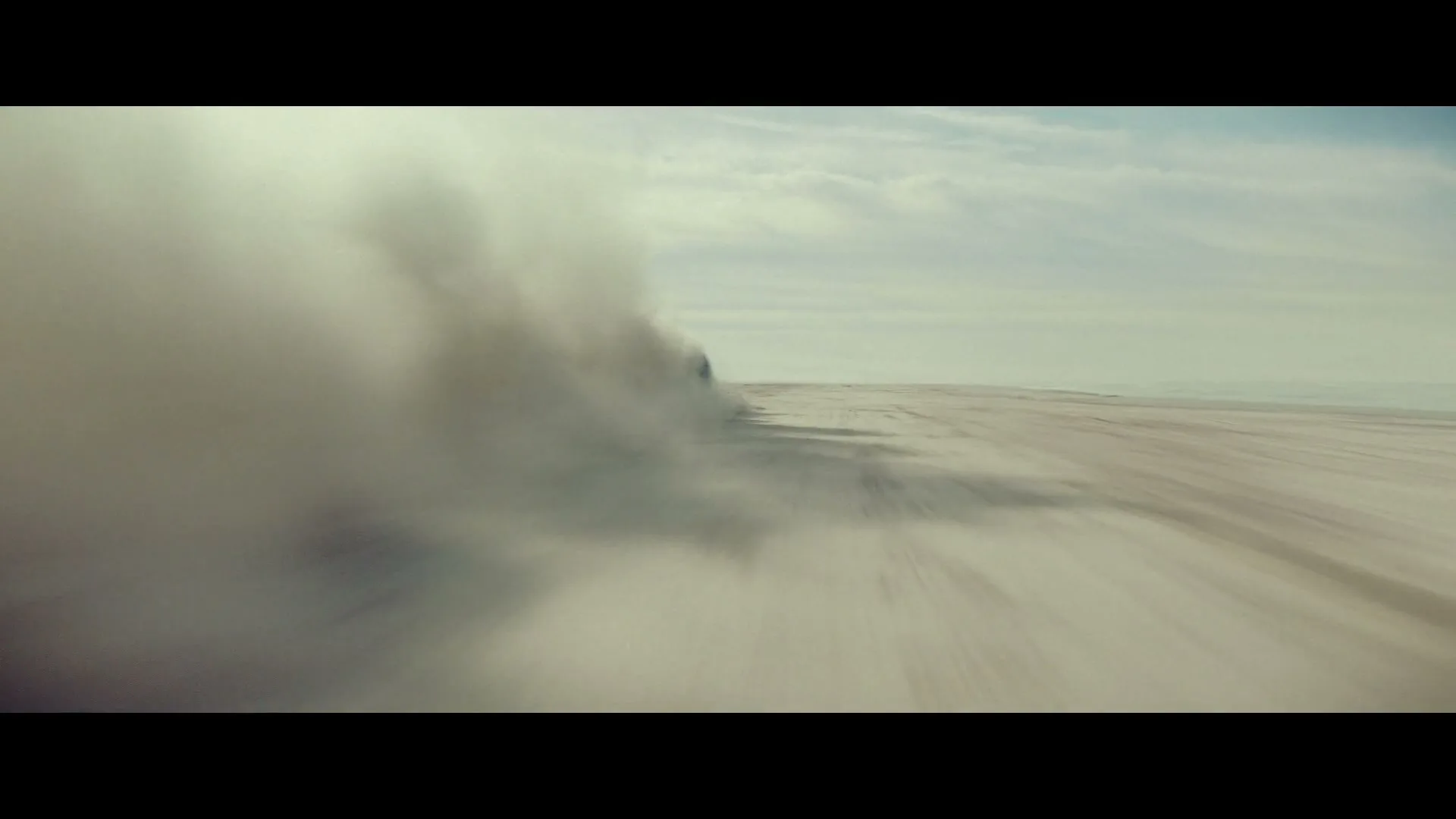 A wide shot shows a vast, flat, dusty landscape under a cloudy sky, with a vehicle speeding past, kicking up a large cloud of dust.