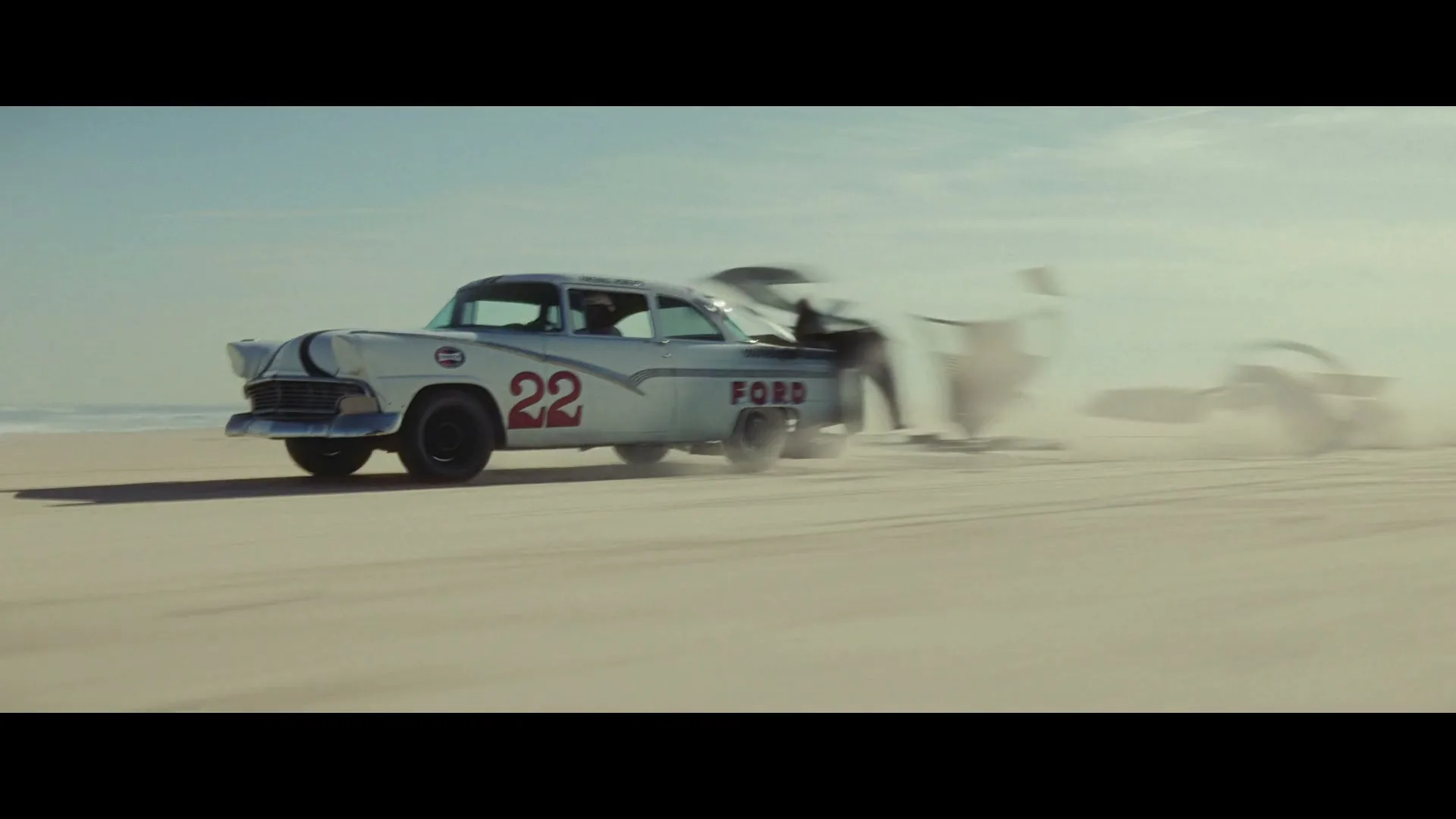 A vintage white Ford race car with the number 22 on its side drives on a sandy beach, kicking up dust as it moves.