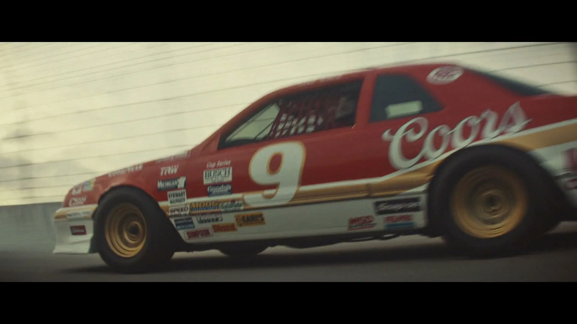 A red and white stock car with the number 9 and "Coors" sponsorship speeds around a race track under an overcast sky.