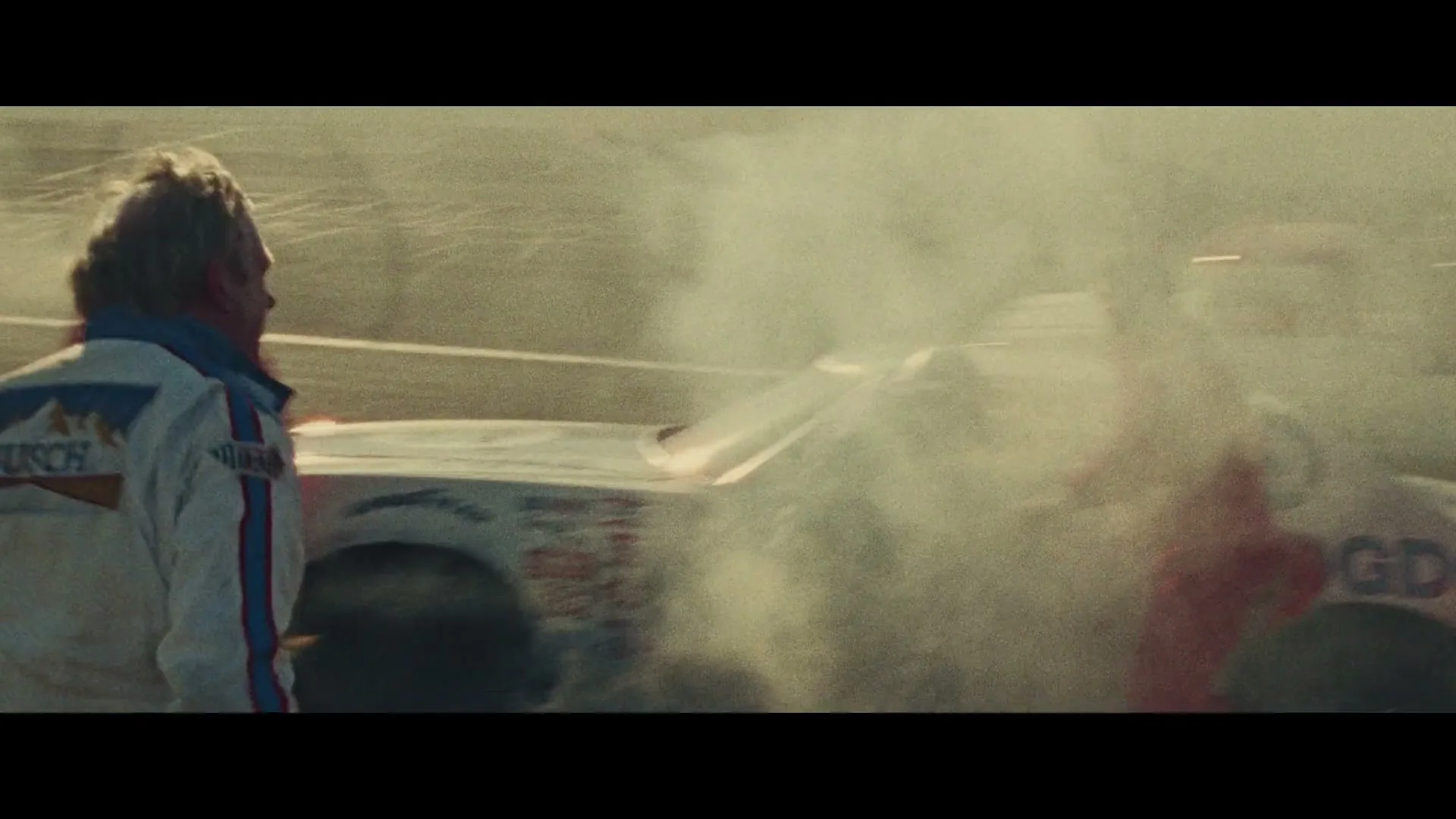 A race car driver in a white and blue jacket watches as a pit crew member in a red suit extinguishes a smoking race car on a track.