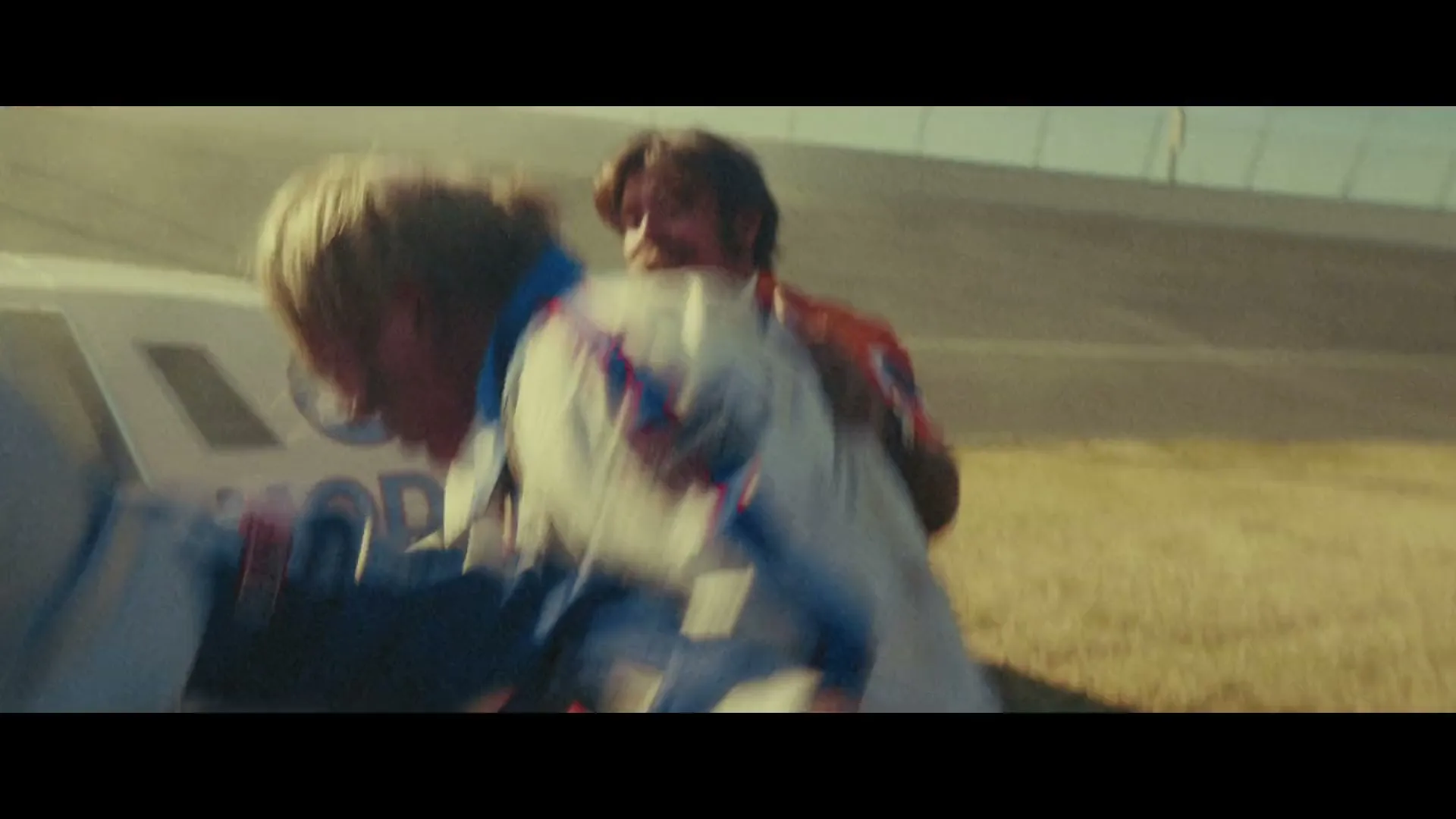 Two male pit crew members in orange uniforms help a male race car driver in a white and blue uniform out of a crashed car on a race track.