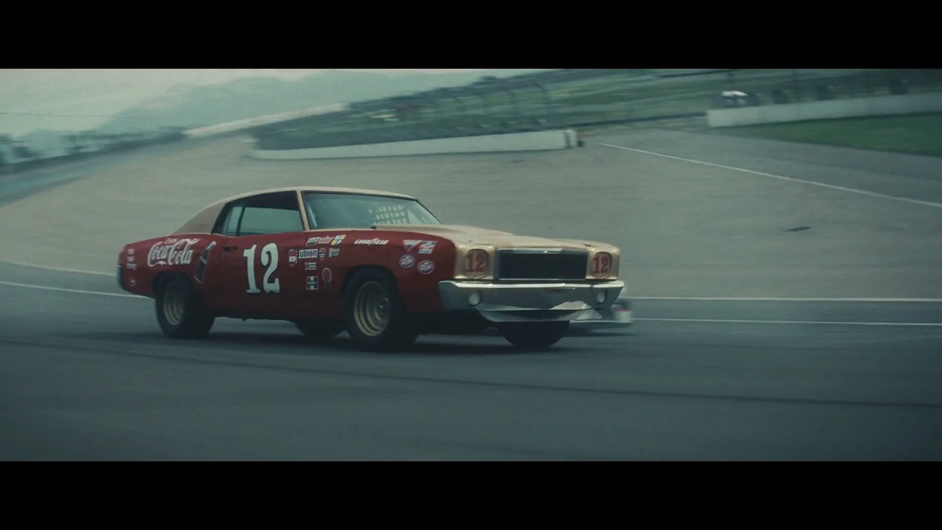 A vintage red and gold race car with the number 12 and Coca-Cola logo drives on a race track, followed by a modern black race car.
