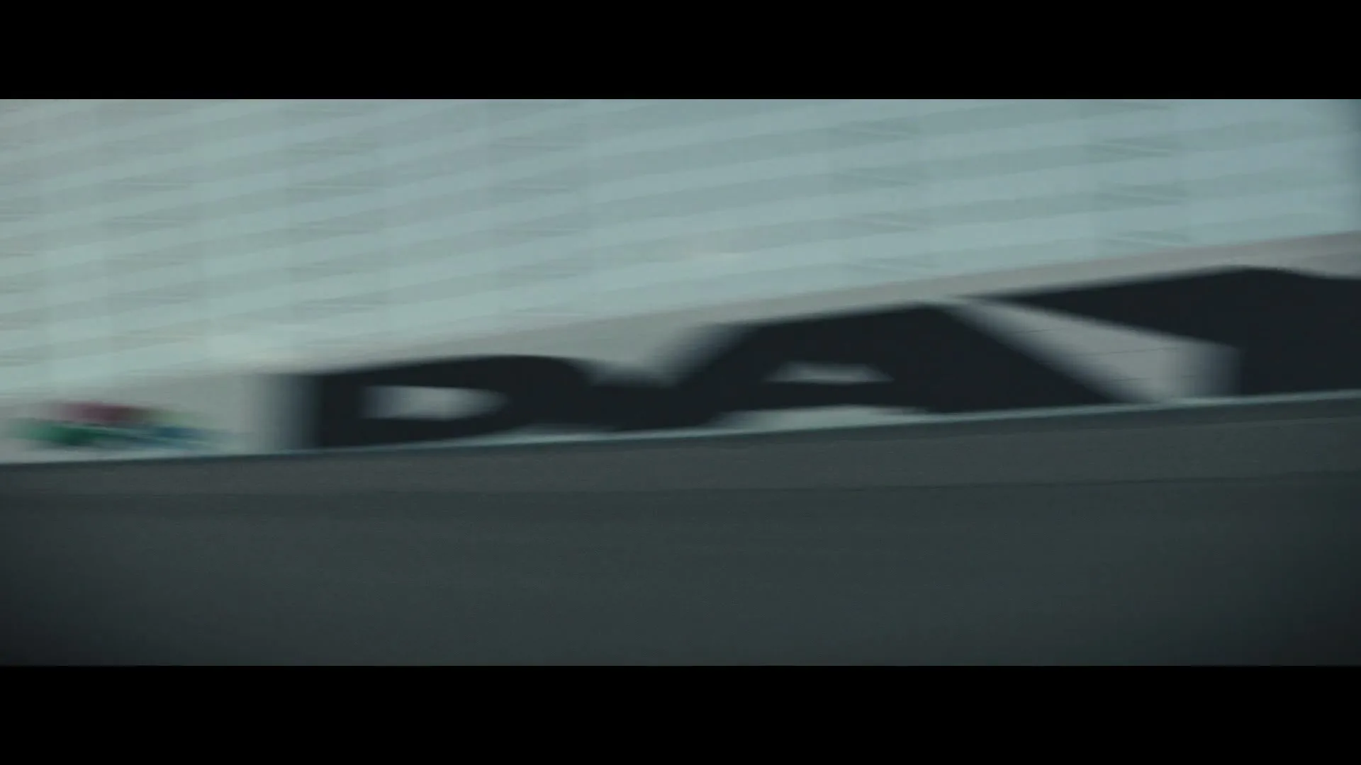 A blurred shot of a race track wall with the word "DAYTONA" visible, as a dark race car speeds by.