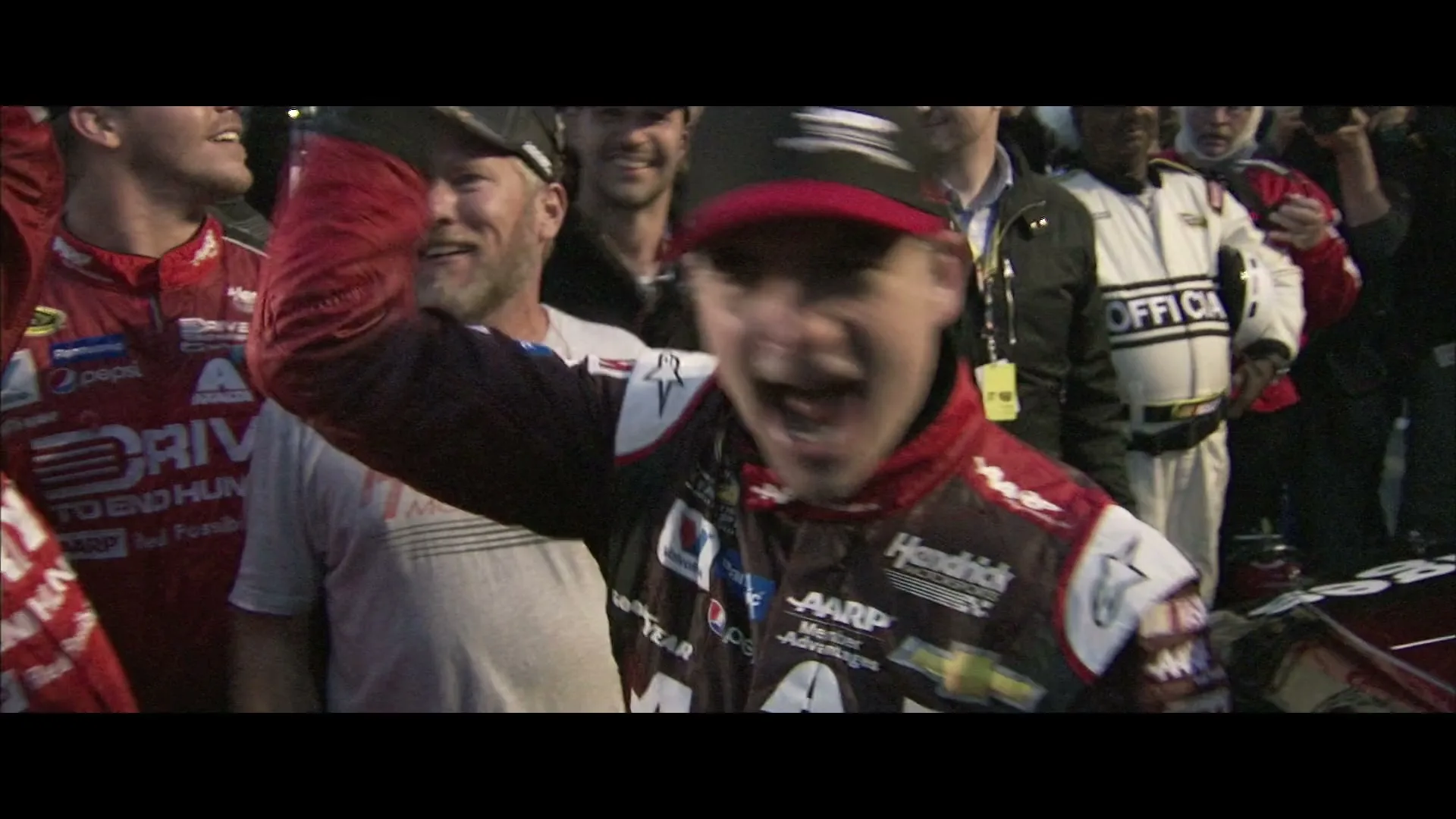 A male race car driver in a red and black racing suit and cap cheers with his arm raised, surrounded by other men in a celebratory moment at a race track.