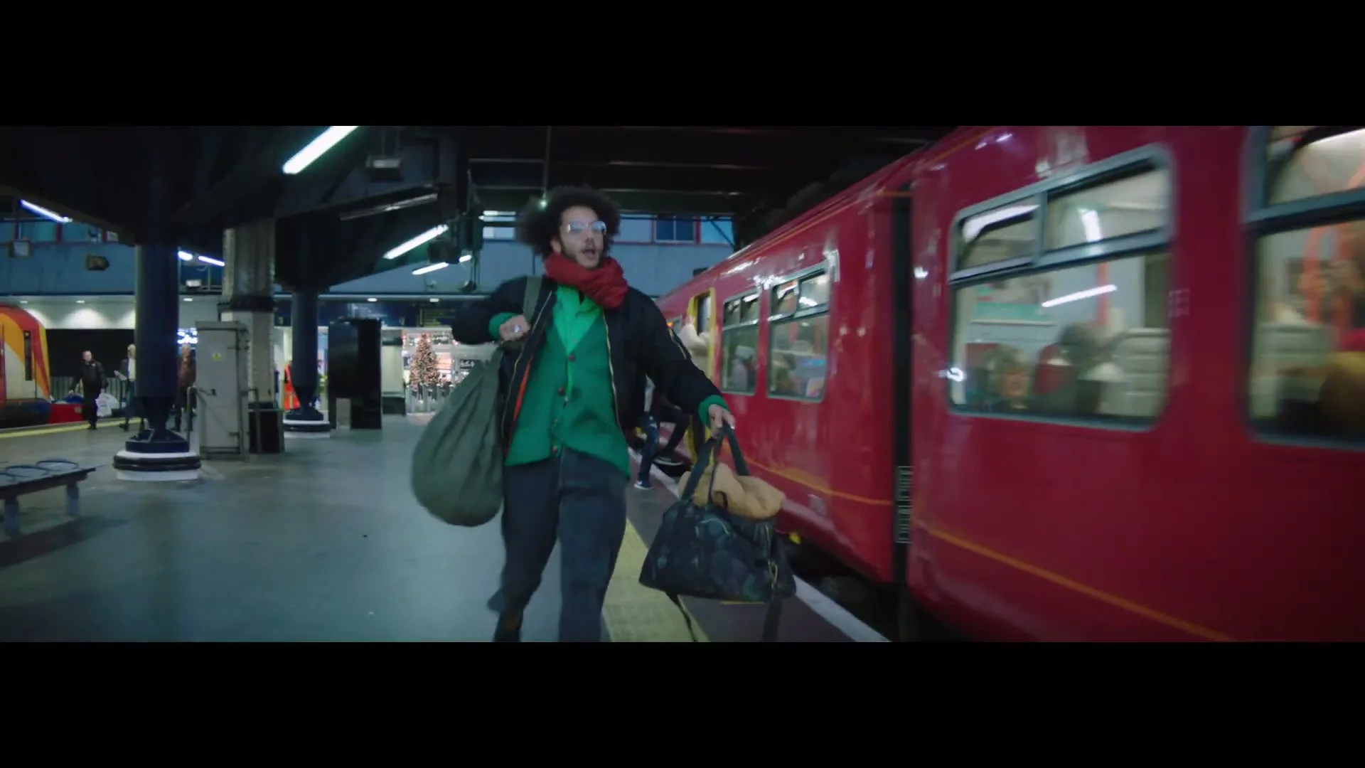A young man with curly hair, wearing a green shirt and red scarf, runs alongside a red train on a station platform, carrying multiple bags.
