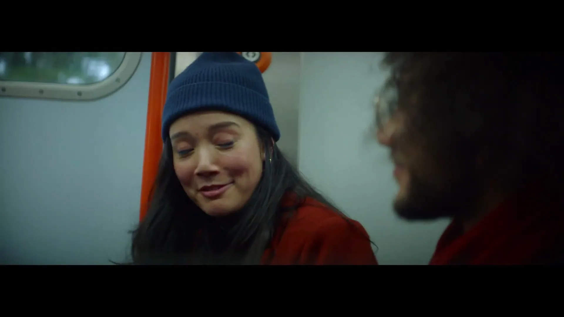 A young woman in a blue beanie and red coat smiles and talks to a man on a train, looking surprised.