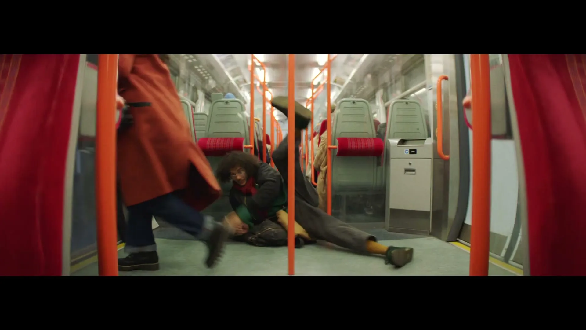 A young man with curly hair falls to the floor of a moving train, while another person walks past him.