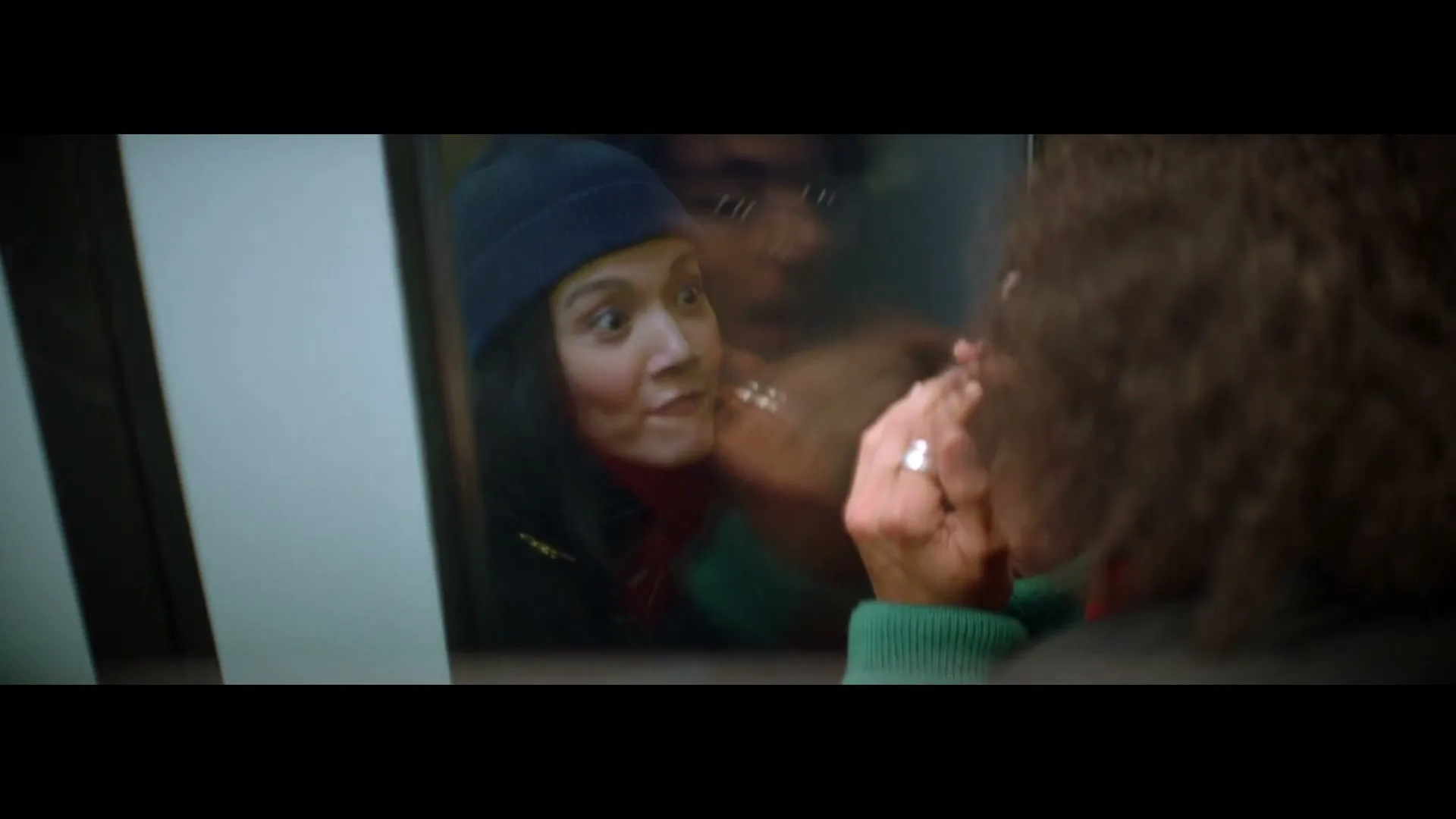 A young woman with a blue beanie and red scarf looks surprised through a window at a person with curly hair, who is out of focus.