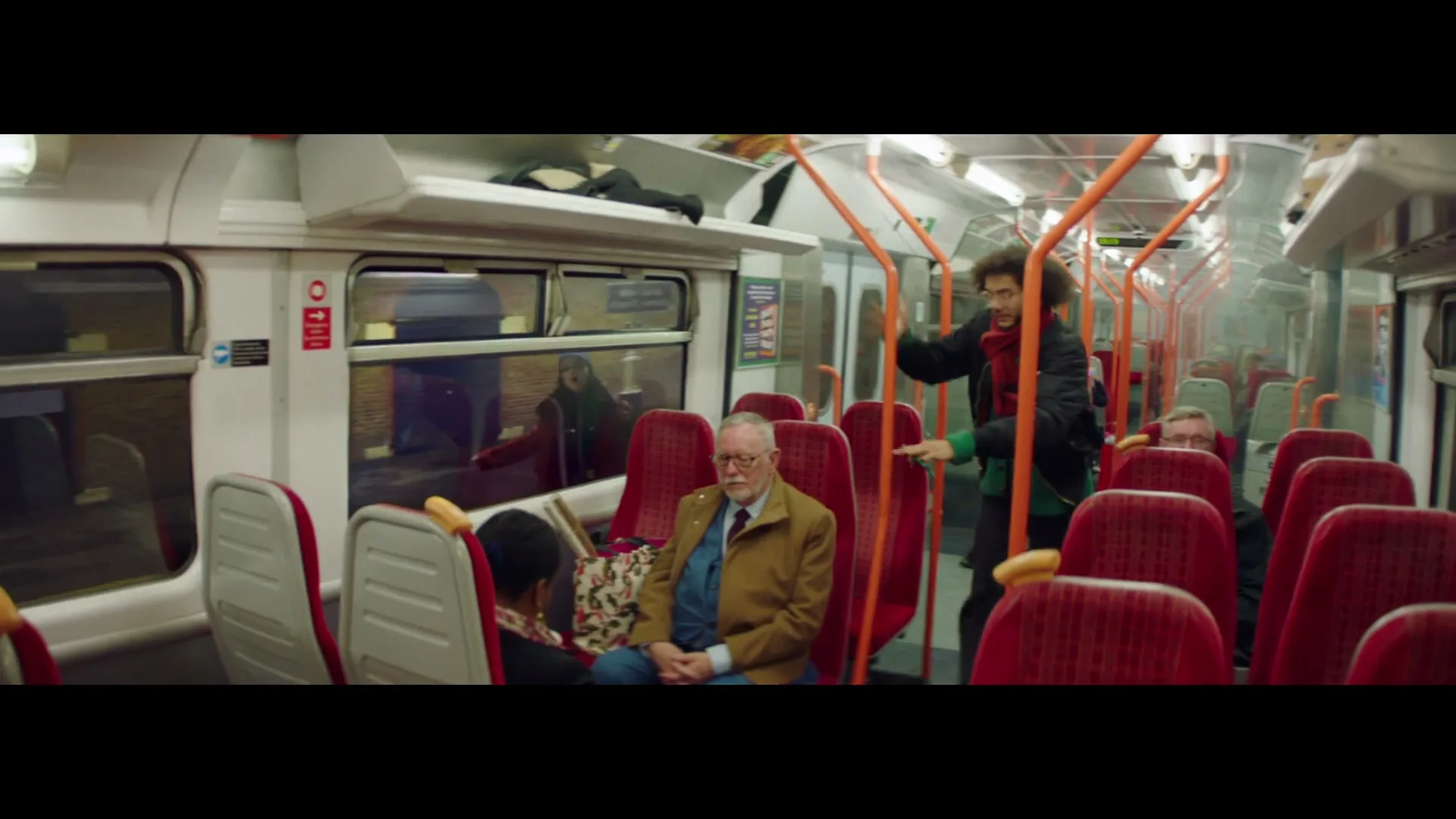 A young man with curly hair and a red scarf walks down the aisle of a train, looking at passengers, while an older man sits in the foreground.