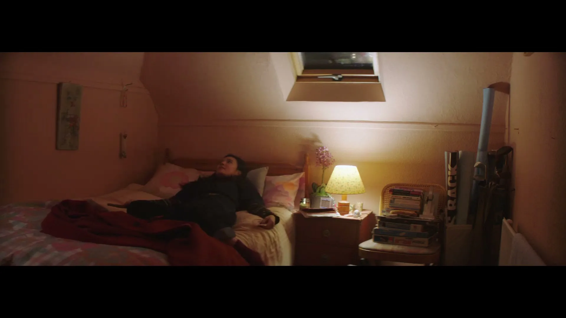A young woman with long dark hair, wearing dark clothes, falls back onto her bed in a cozy attic bedroom, looking at her phone.