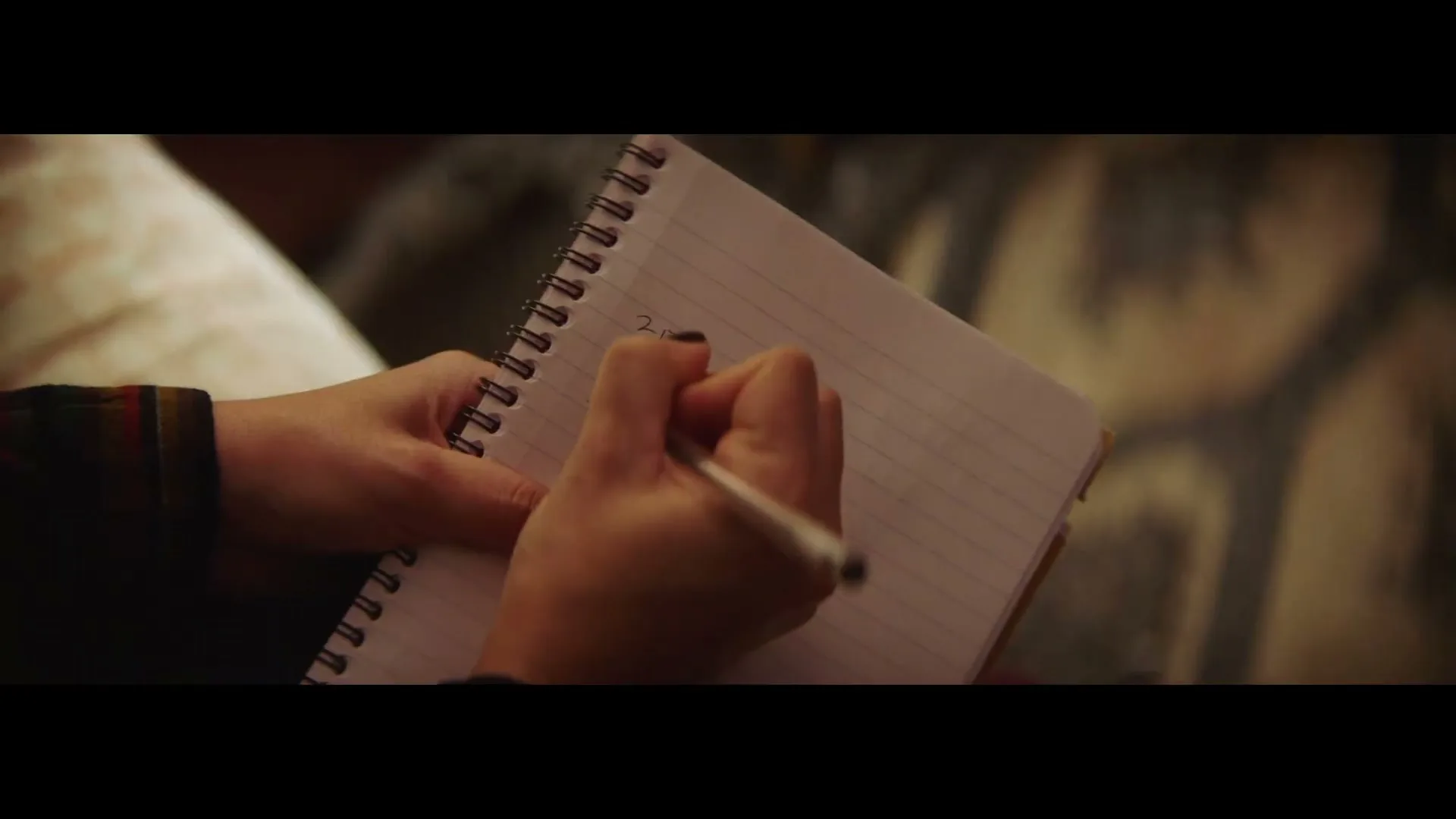A person's hands write numbers in a spiral notebook with a pen, seen from a high angle in an interior setting.
